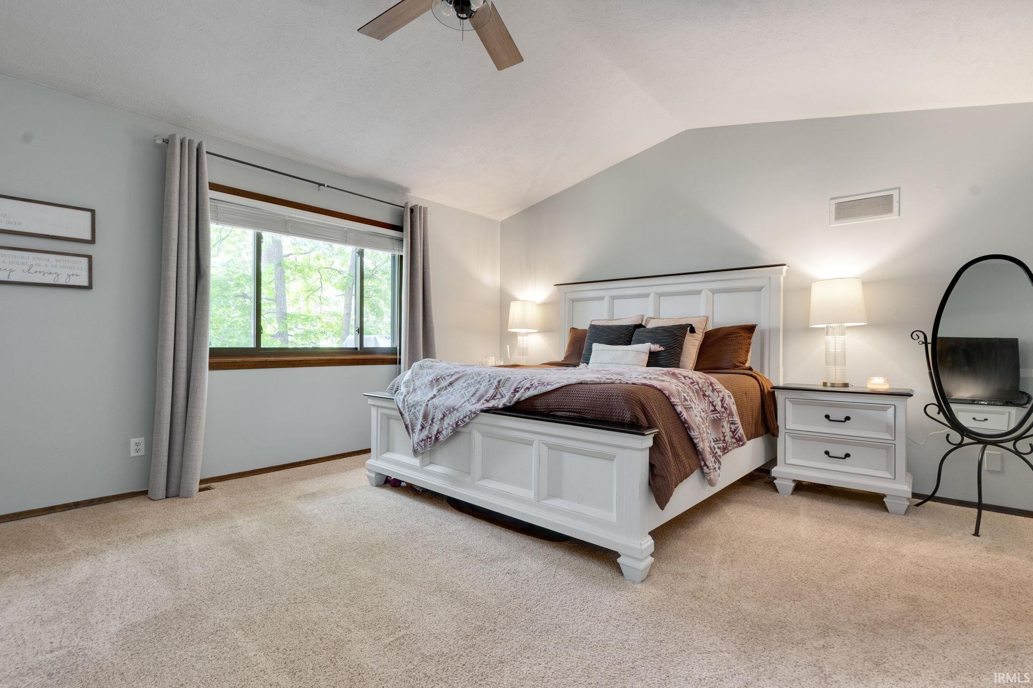 Vaulted ceiling bedroom featuring plush carpet flooring, a large window with wood trim, and a ceiling fan