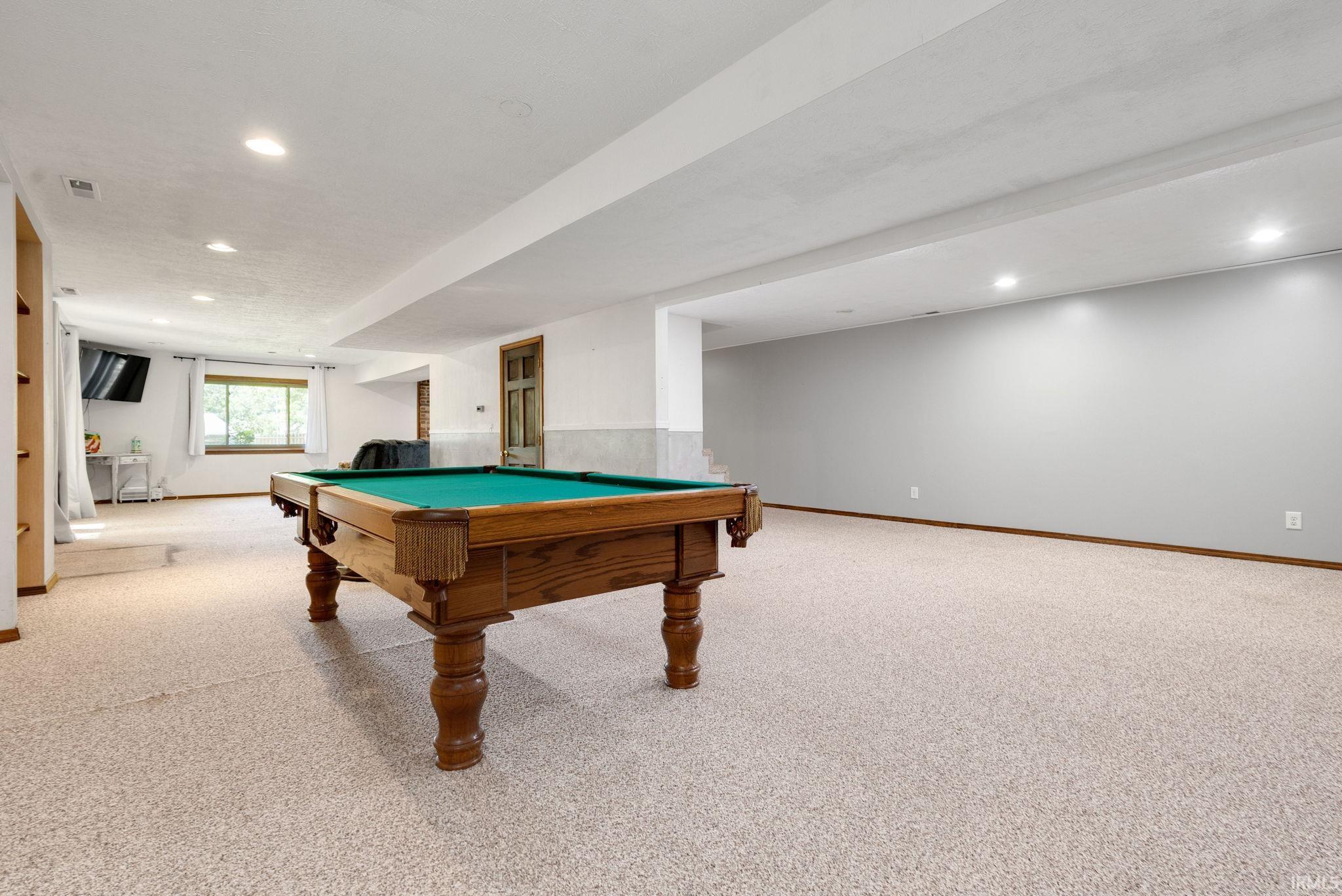 Spacious recreational room featuring light-colored carpeting, recessed lighting, and a large window providing ample natural illumination