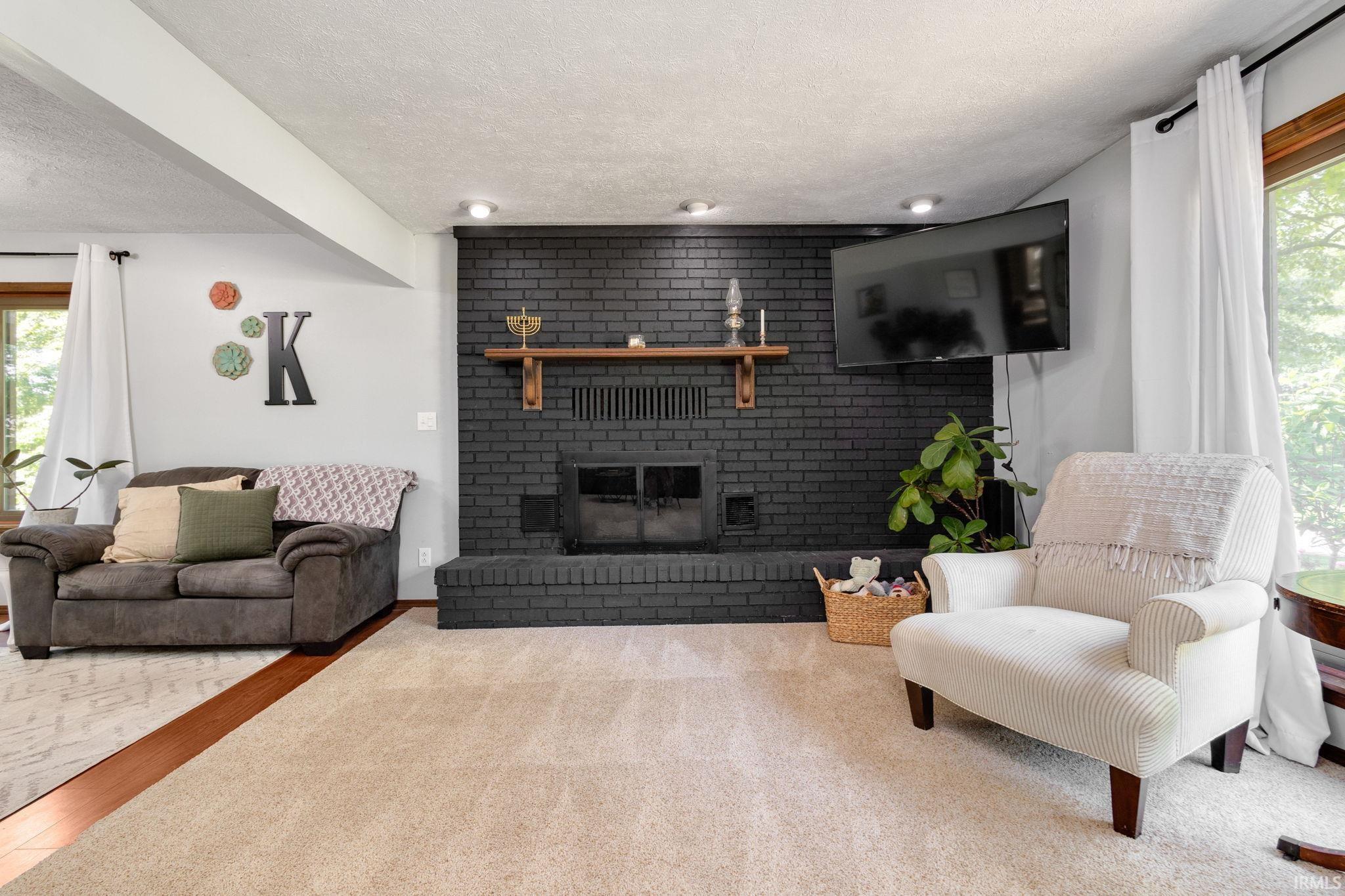 Spacious living area featuring a painted brick fireplace with a wood mantel, recessed lighting, and a window with white curtains