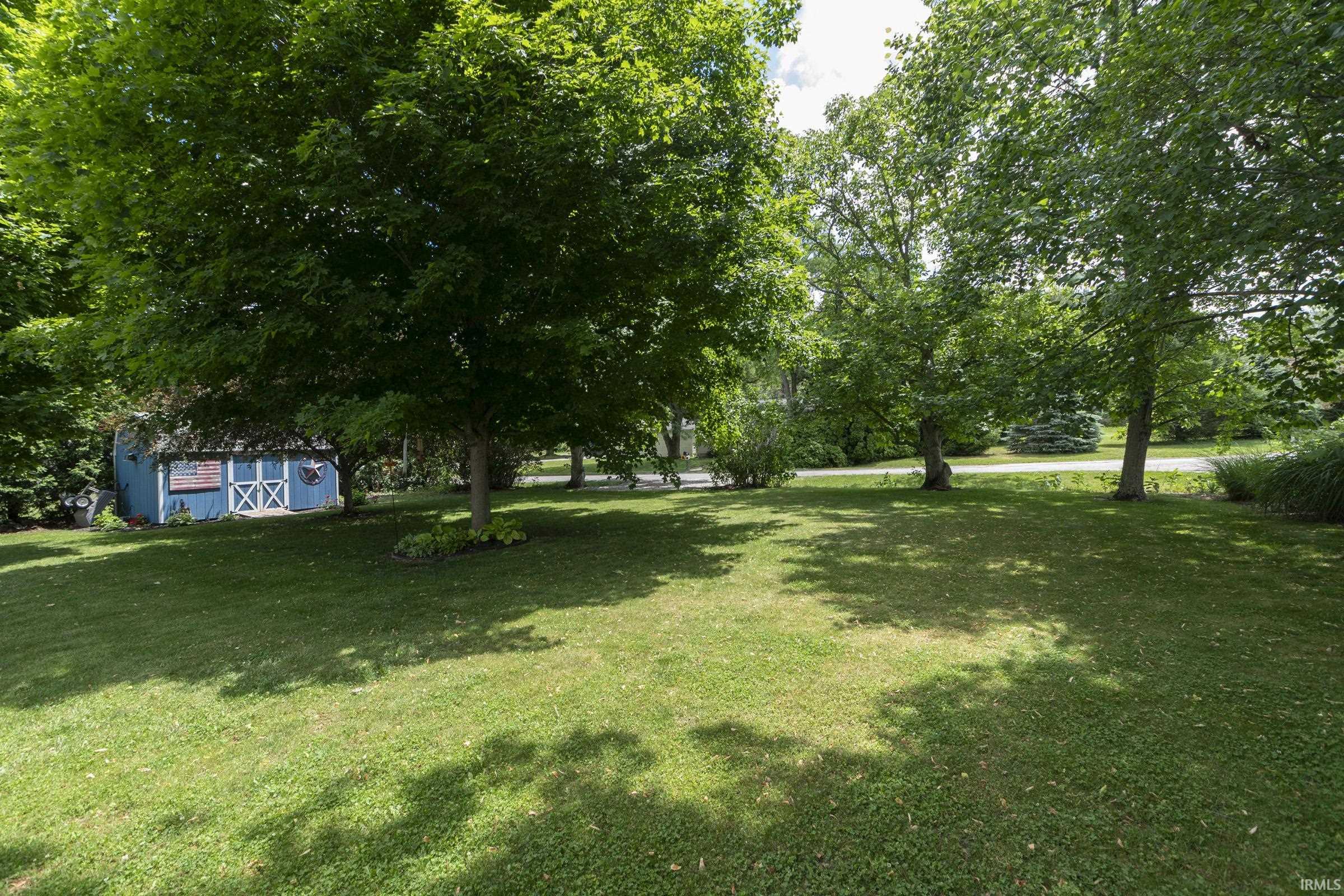 Spacious lawn featuring mature trees providing ample shade, established landscaping, and a blue shed with white trim
