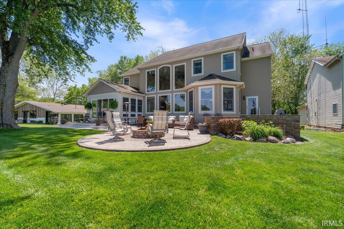 Expansive green lawn featuring a circular paver patio with a built-in fire pit, stone retaining wall with landscaping, and a multi-level tan exterior home with numerous windows