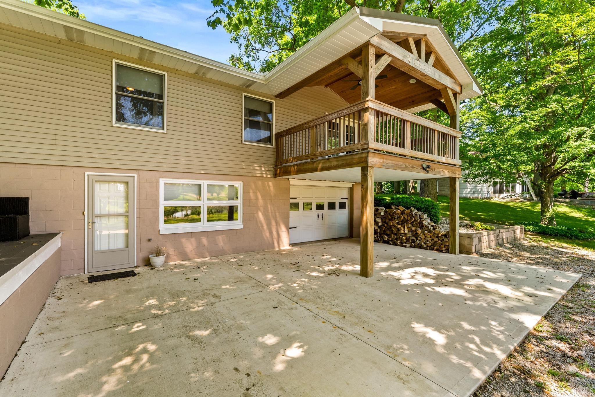 Rear exterior featuring a covered porch with natural wood railing and exposed beams, a two-story design with beige siding, a ground-level patio, and a single-car garage