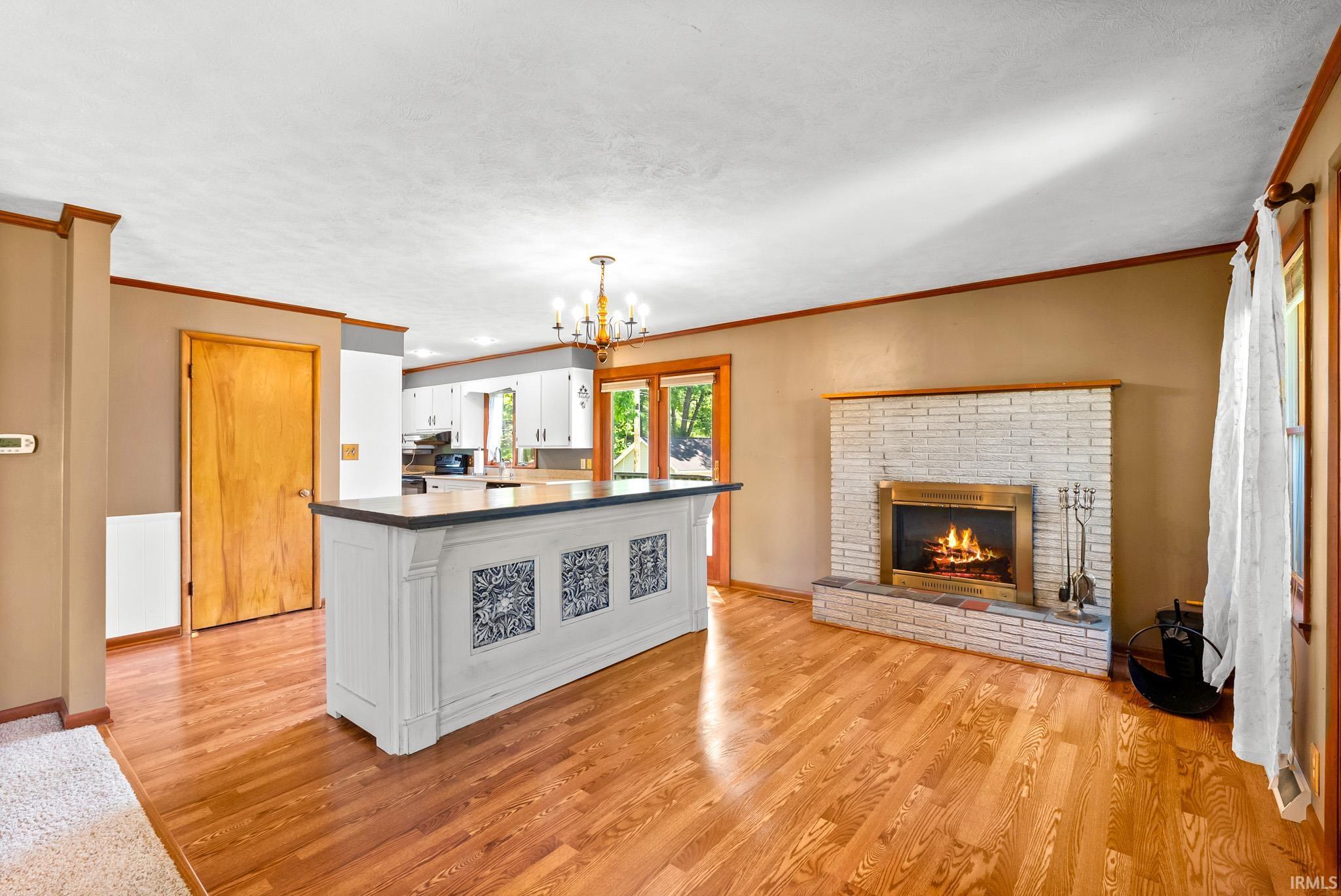 Wood-finish flooring throughout, featuring a white painted brick fireplace with brass firebox, a kitchen island with decorative tile accents and a dark countertop, and an overhead chandelier