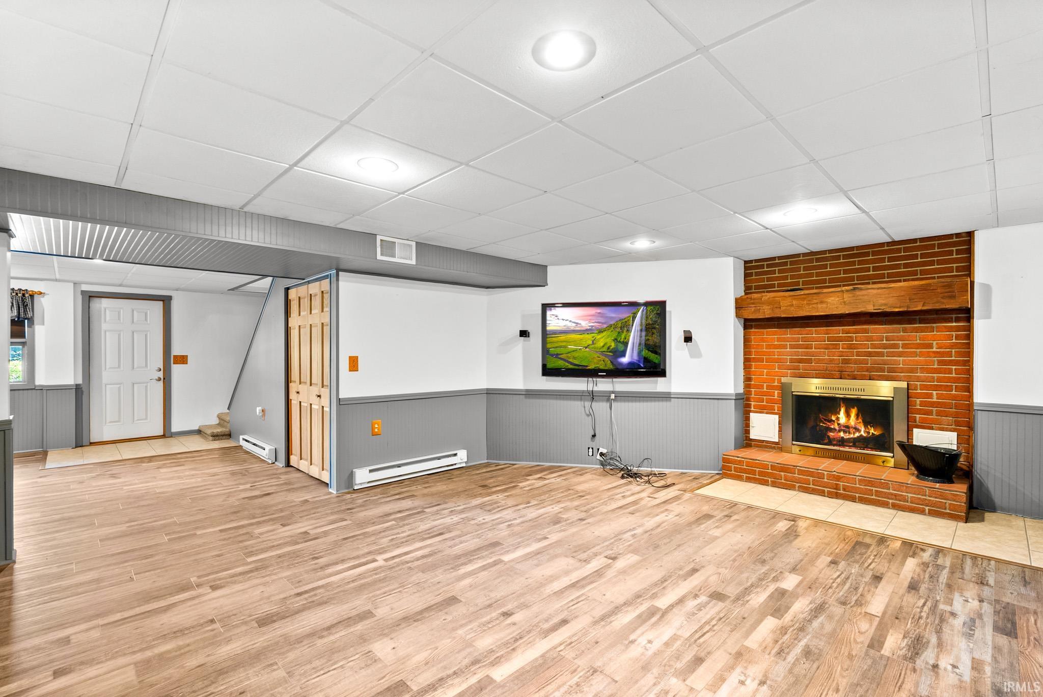 Spacious lower-level room featuring wood-finish flooring, a brick fireplace with a wood mantle, and beadboard wainscoting
