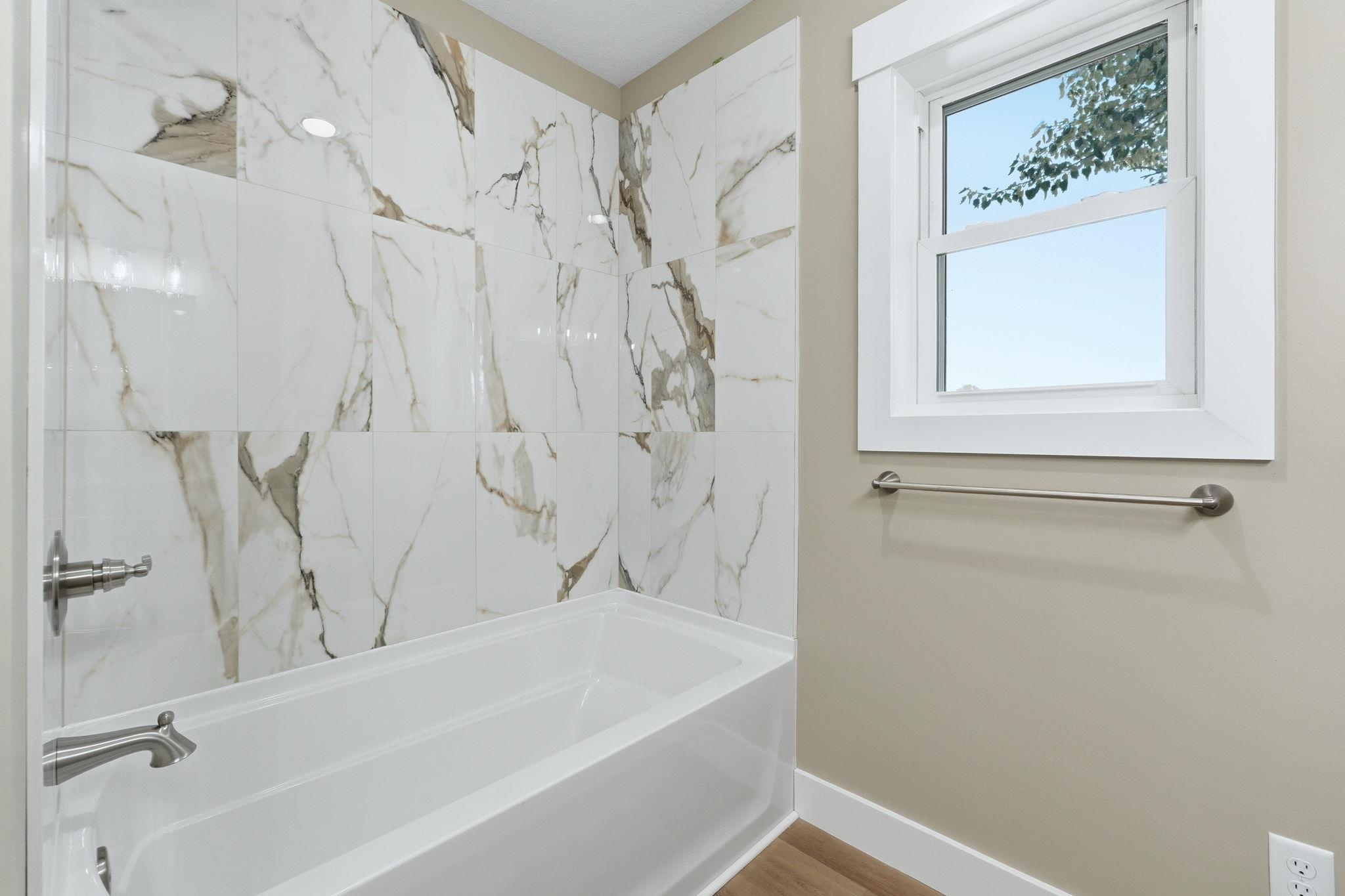 Bathtub with white and gray veined wall tile surround, brushed nickel fixtures, and a white framed window