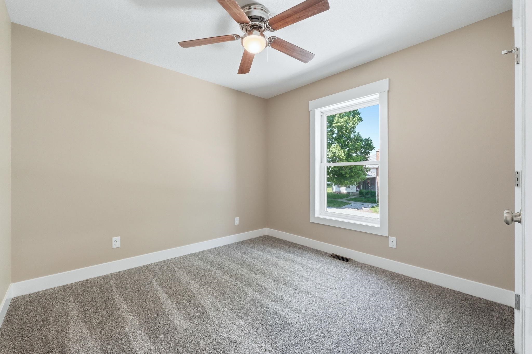 Neutral-toned bedroom featuring a ceiling fan with wood-finish blades, a framed window, light gray carpeting, and white baseboards