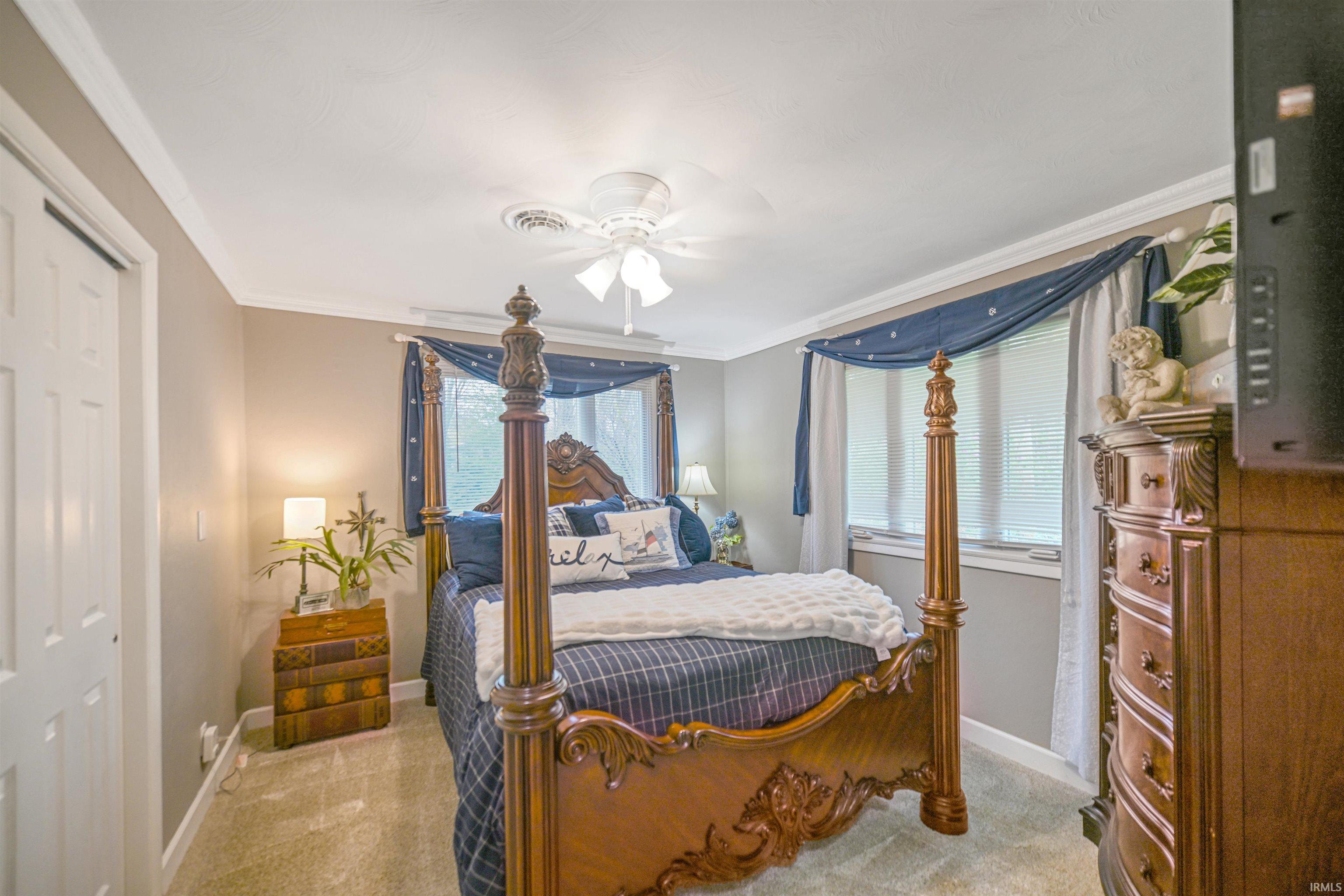 Bedroom featuring light neutral wall paint, white crown molding, carpet flooring, a ceiling fan with light fixture, and windows with blinds