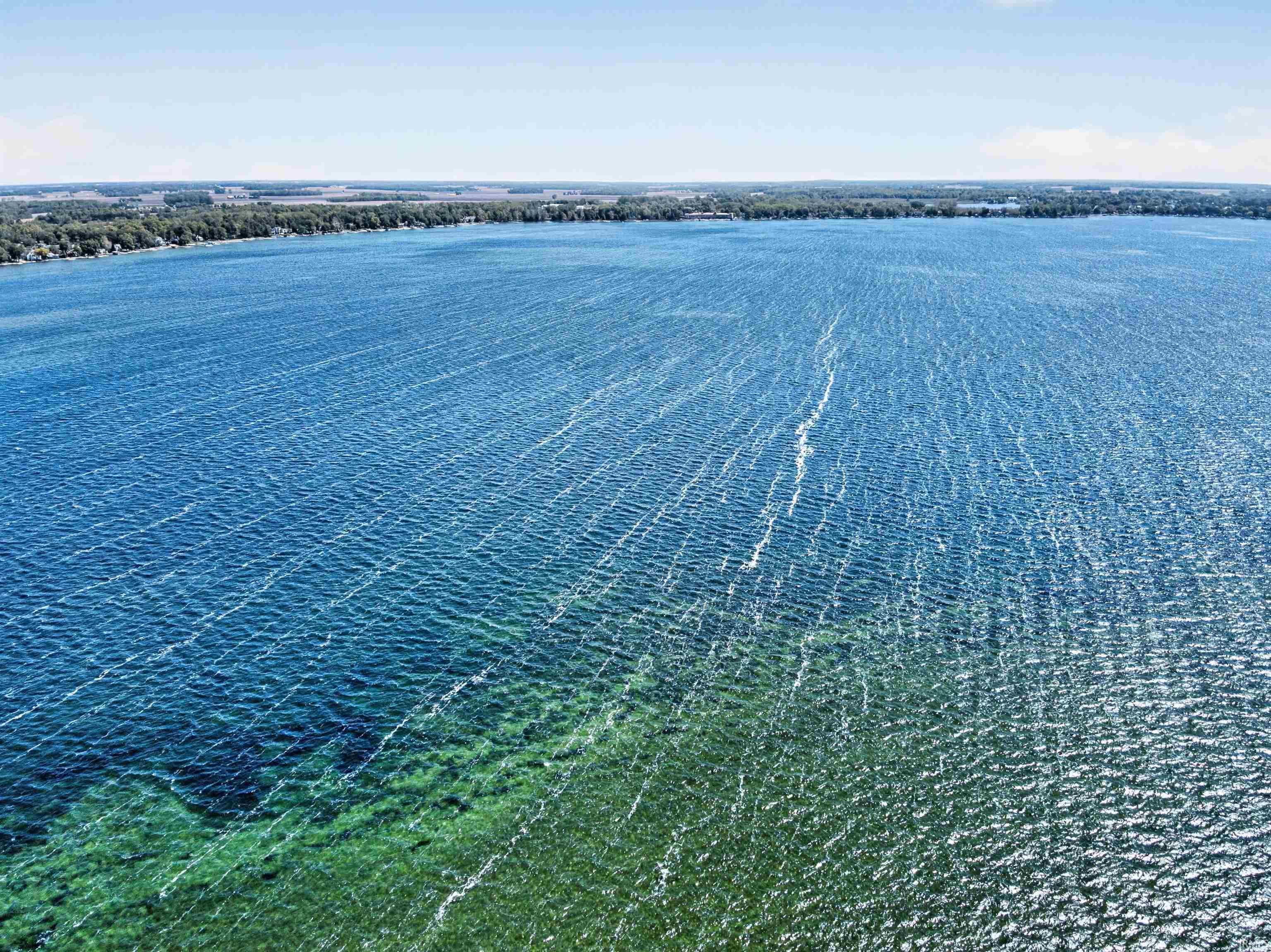 Expansive body of water with a clear shoreline, featuring varied water depths, a distant tree line, and a bright, open sky