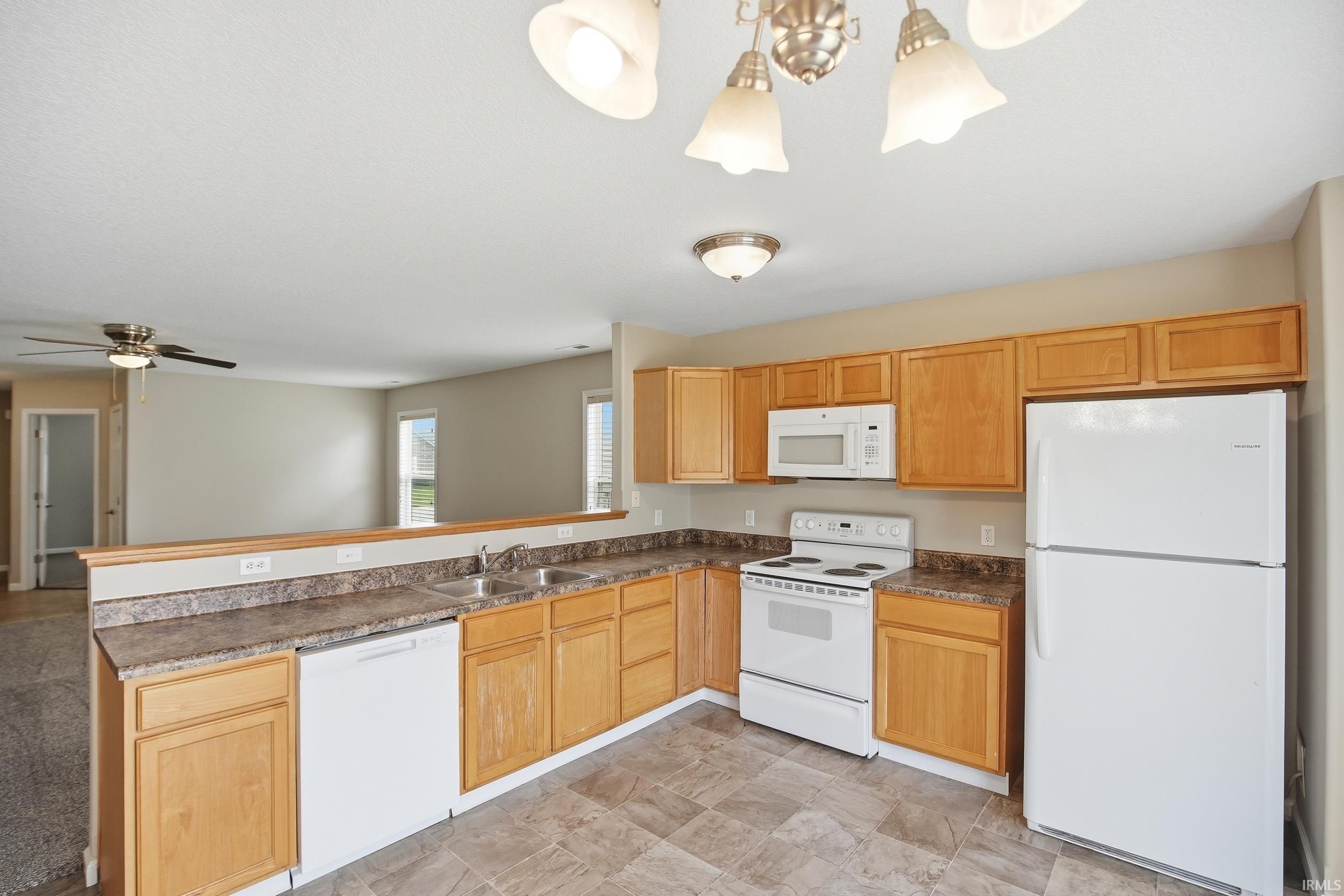 Kitchen featuring light wood cabinetry, laminate countertops, a double basin stainless steel sink, and white appliances including a refrigerator, range, microwave, and dishwasher