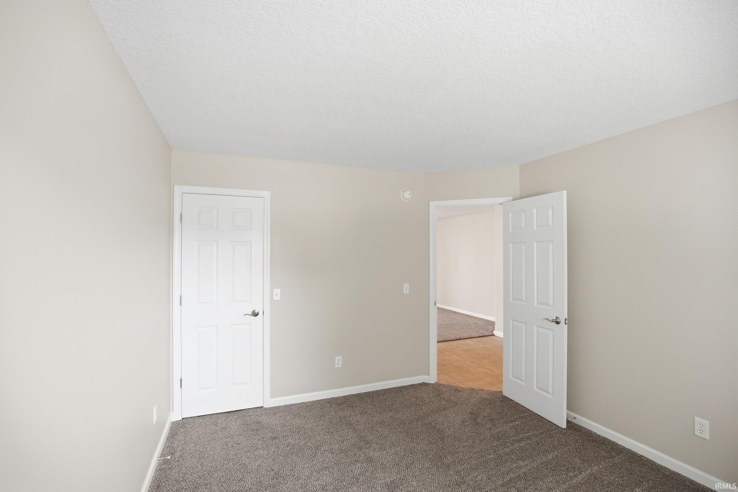 Interior room featuring neutral wall paint, dark grey carpeting, a white paneled door with brushed nickel hardware, and textured white ceilings