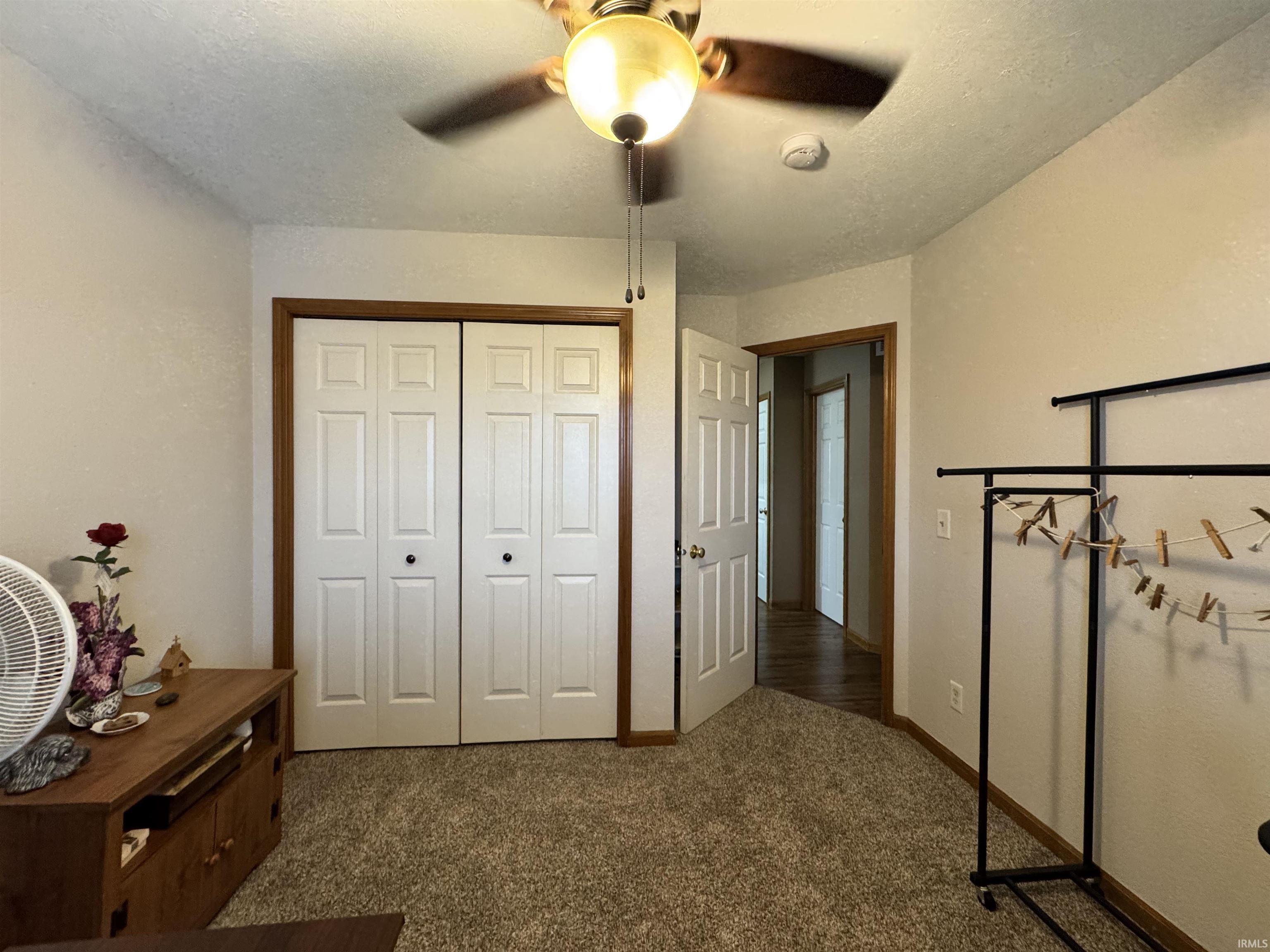 Carpeted room featuring a ceiling fan with integrated lighting, bi-fold closet doors with wood trim, and a doorway opening to a hall with wood-finish flooring