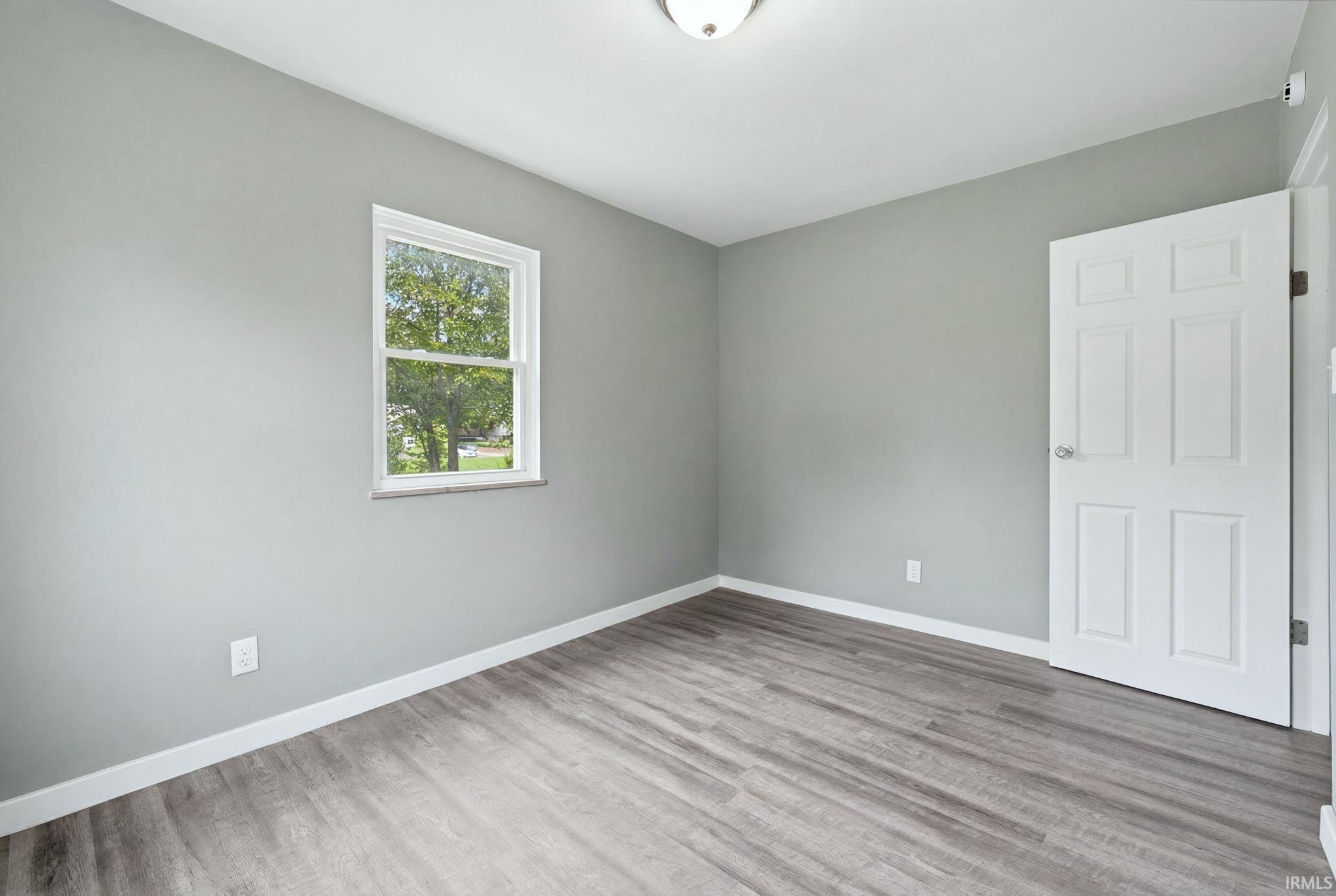 Room featuring wood-finish flooring, light gray walls, white trim, a single window, and a white paneled door