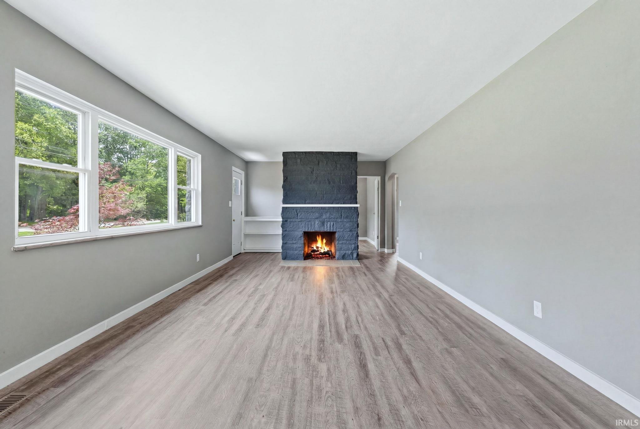 Inviting living area featuring a stone fireplace with a white mantel, wood-finish flooring, and a large window overlooking lush greenery