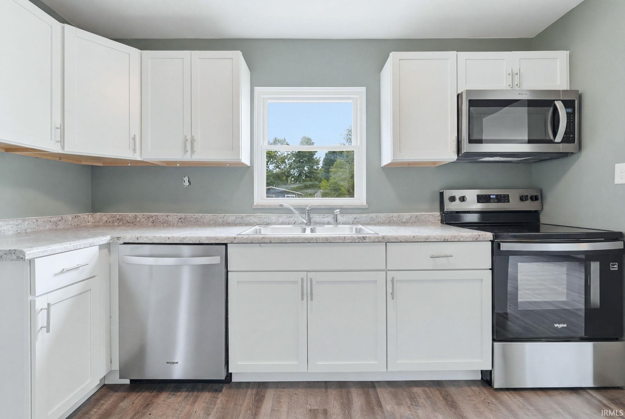 Bright kitchen featuring white cabinetry, light-toned countertops, stainless steel appliances, a double basin sink, and wood-finish flooring