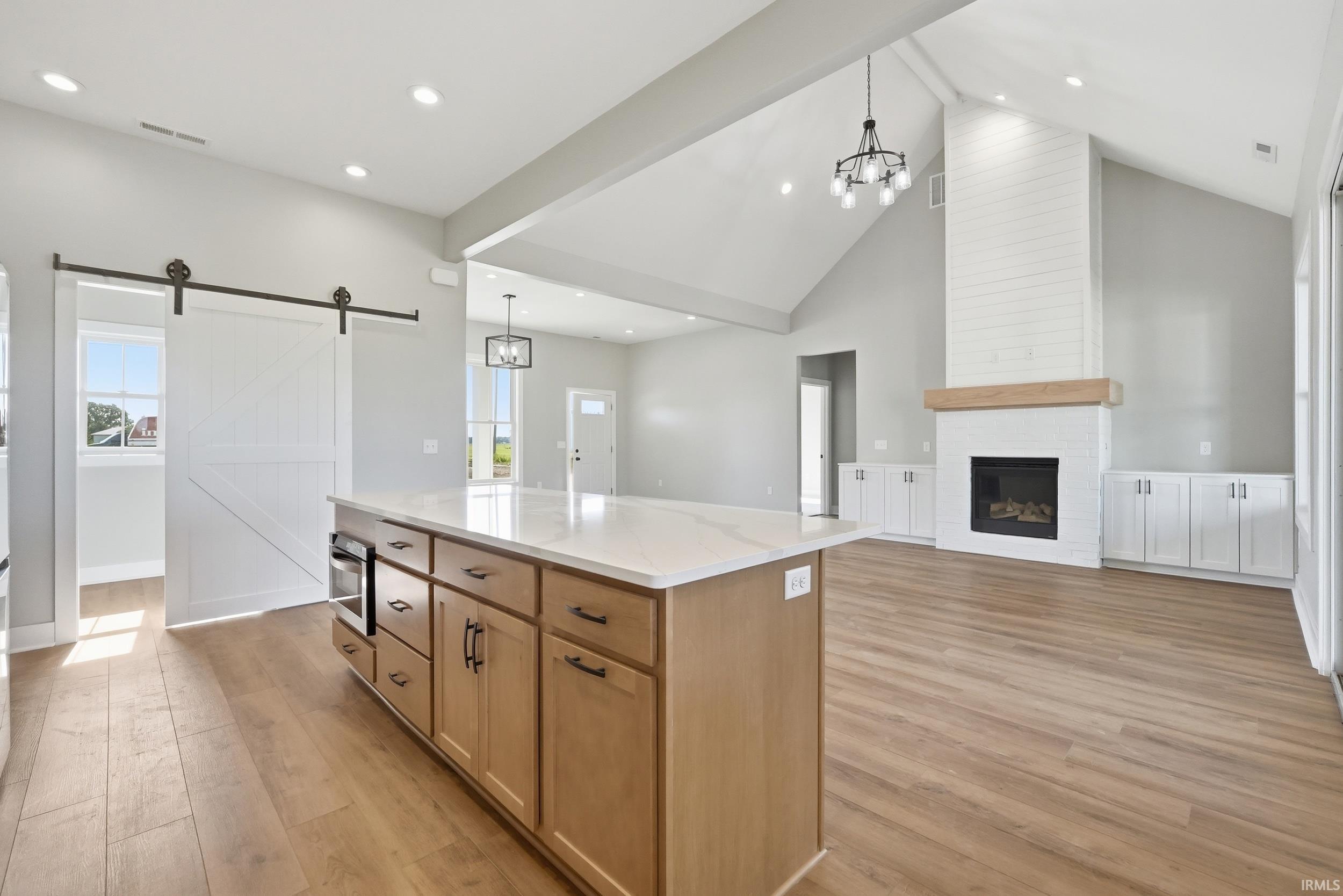 Open-concept living space featuring a central kitchen island with a light-toned countertop, wood-finish flooring, a vaulted ceiling, a white brick fireplace with a wood mantle, and a white barn door
