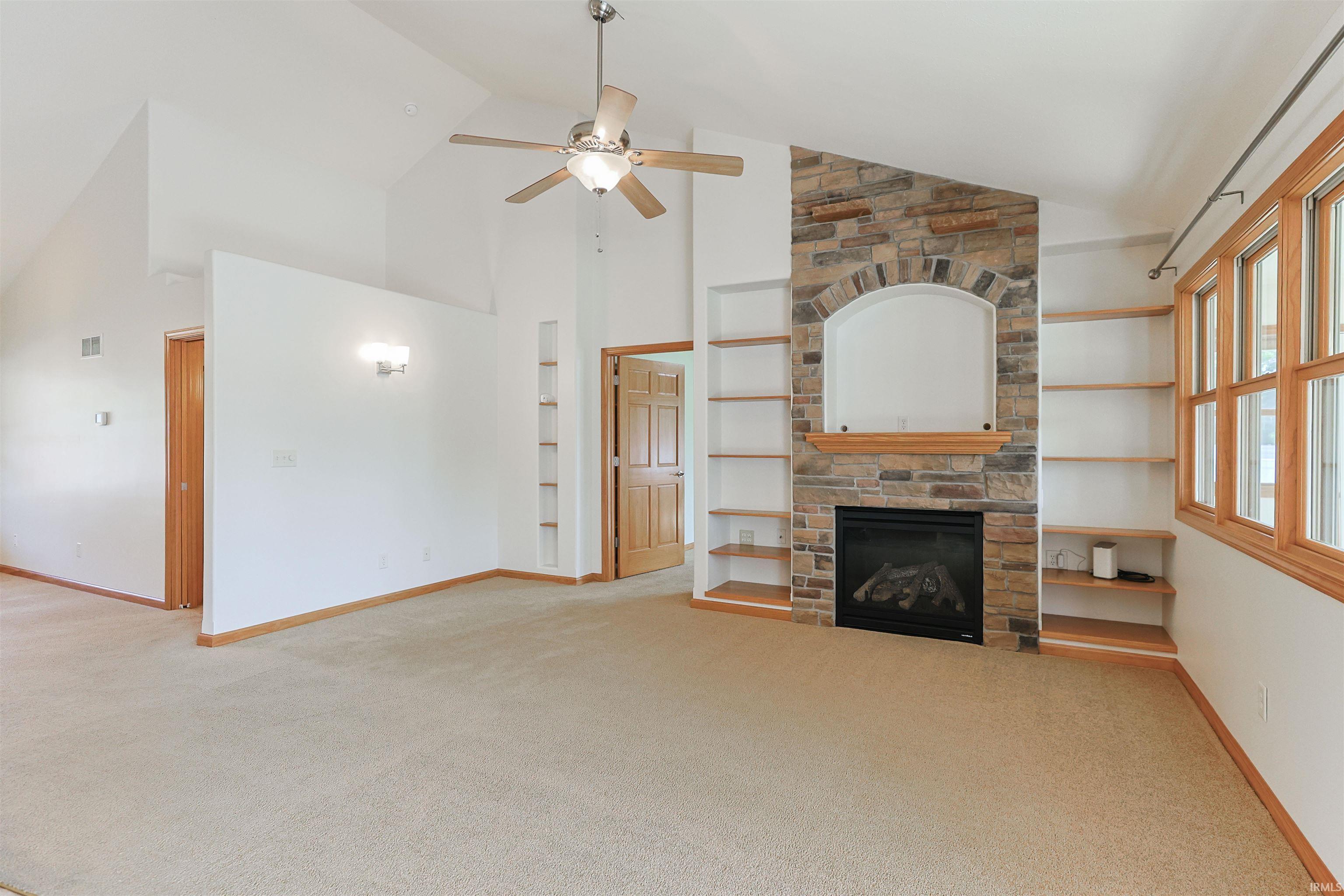 Vaulted ceiling living space featuring a stone-clad fireplace with mantel, built-in shelving, a ceiling fan, and wood-framed windows