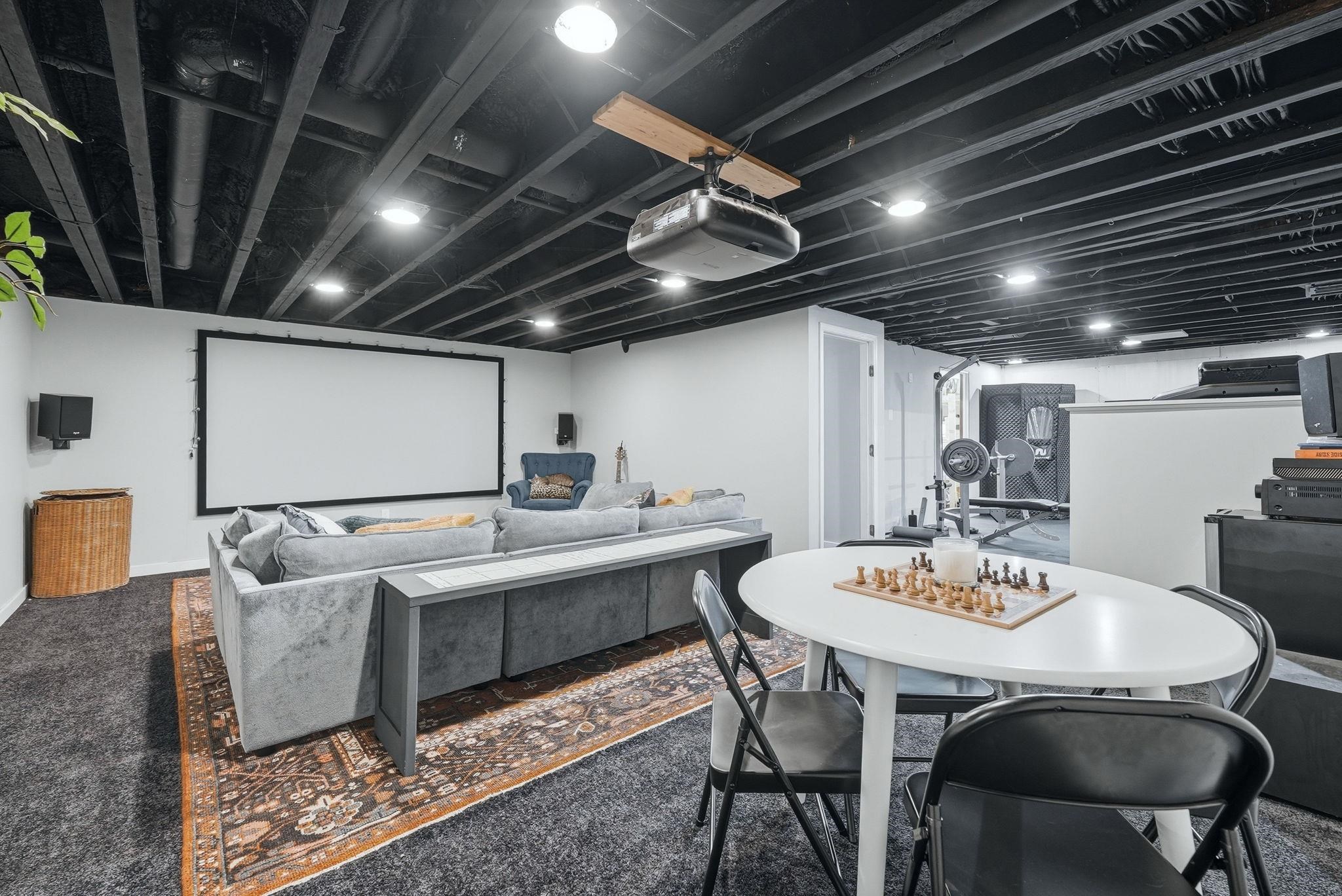 Lower-level entertainment space featuring an exposed beam ceiling with recessed lighting, a large projection screen, and a dedicated fitness area