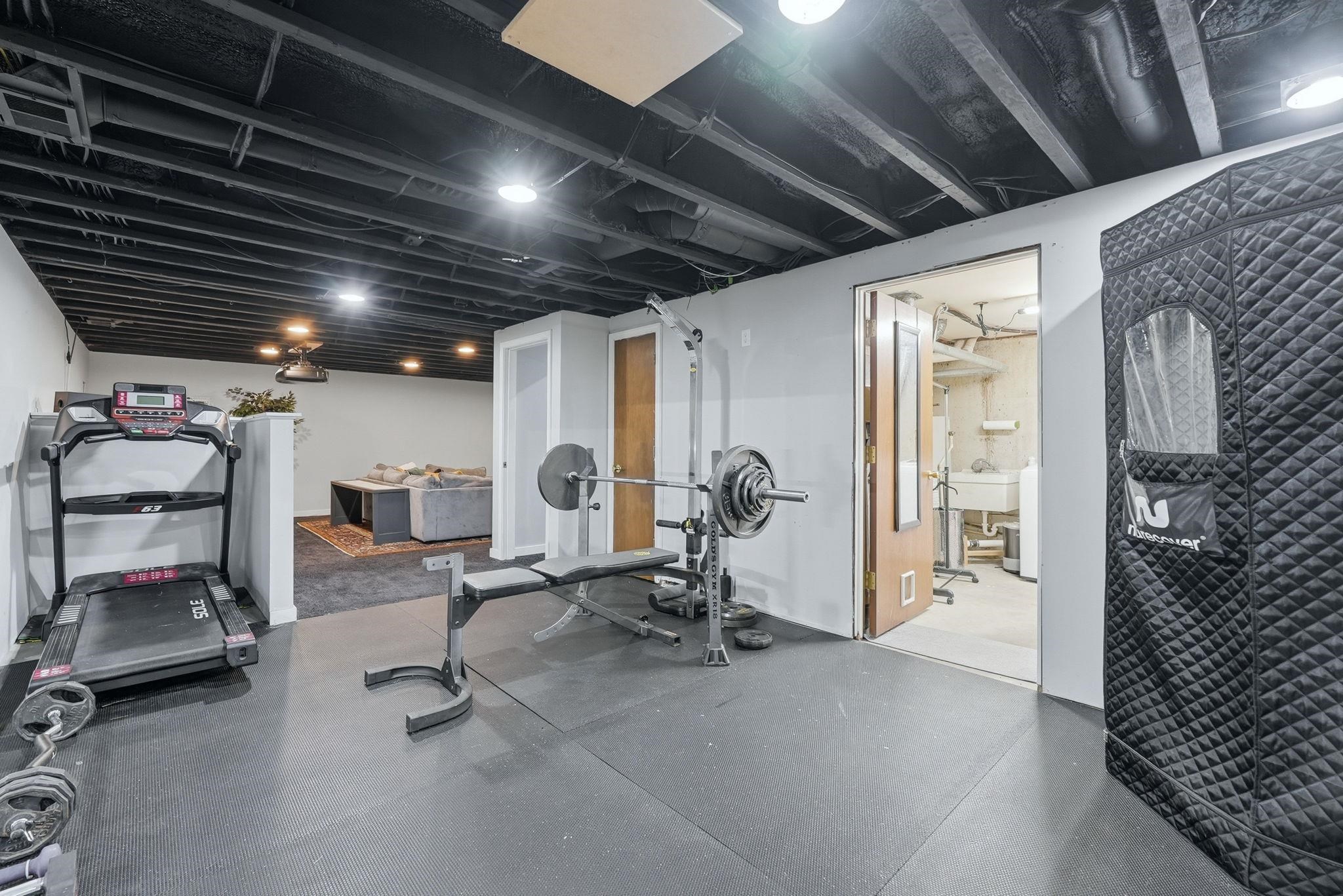Finished basement space featuring exposed painted joist ceiling, recessed lighting, rubber mat flooring, and an additional room beyond