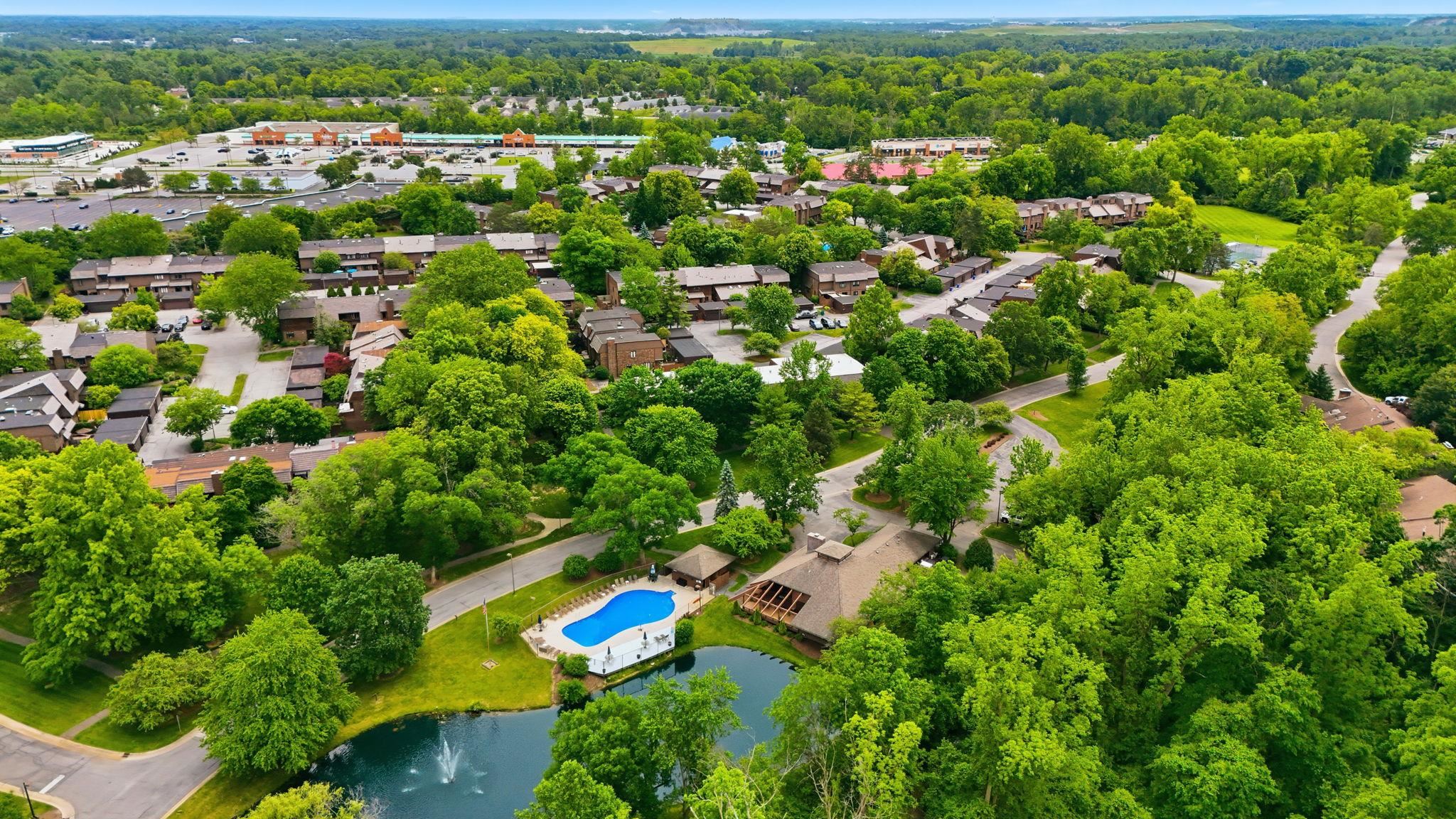 Expansive aerial perspective showcasing a community with mature tree coverage, a sparkling outdoor swimming pool, and a tranquil pond with a water feature