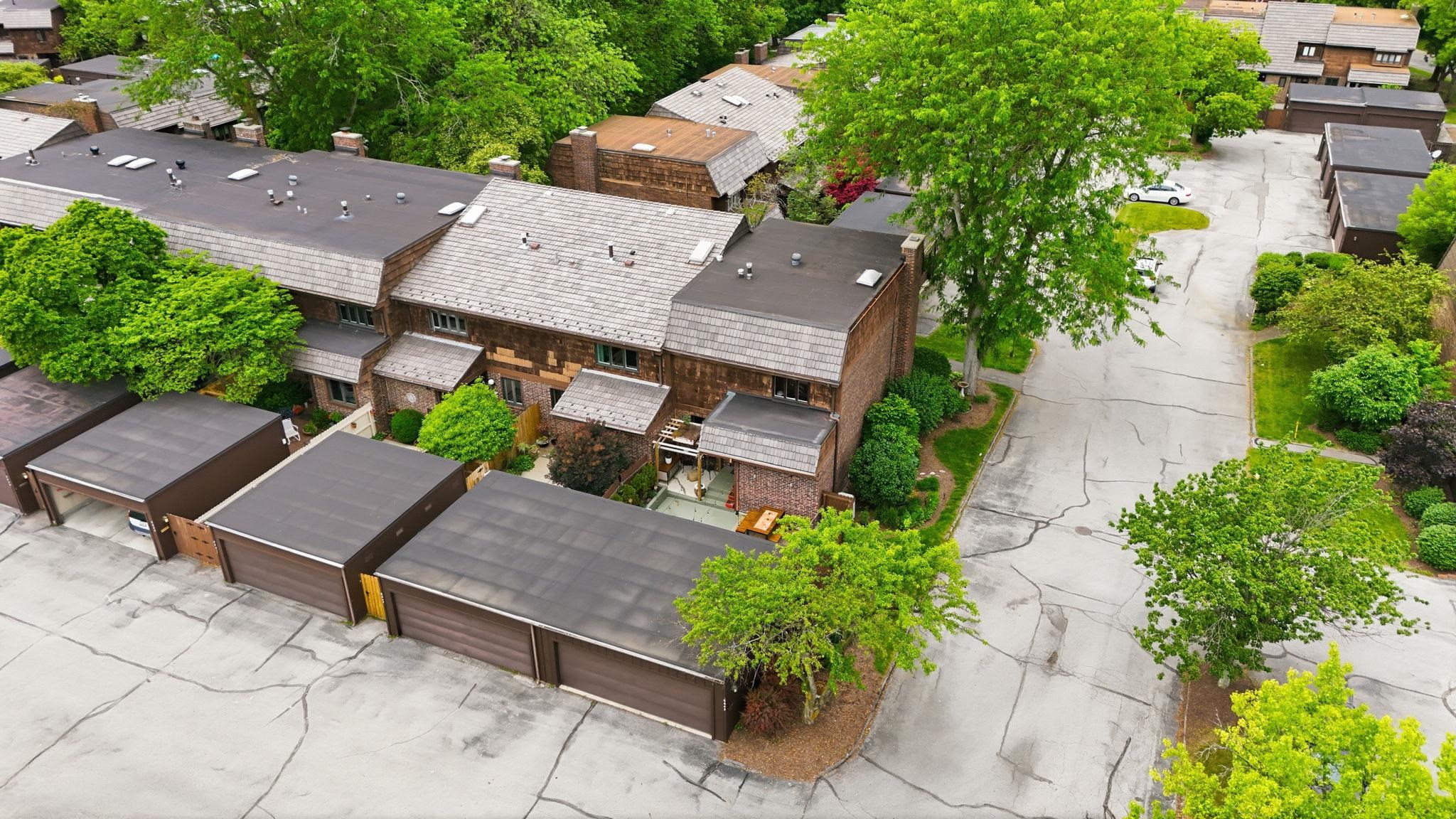 Multi-unit residential property featuring a gray shingle roof, brick and wood-sided exterior, individual garage units with brown doors, and mature trees