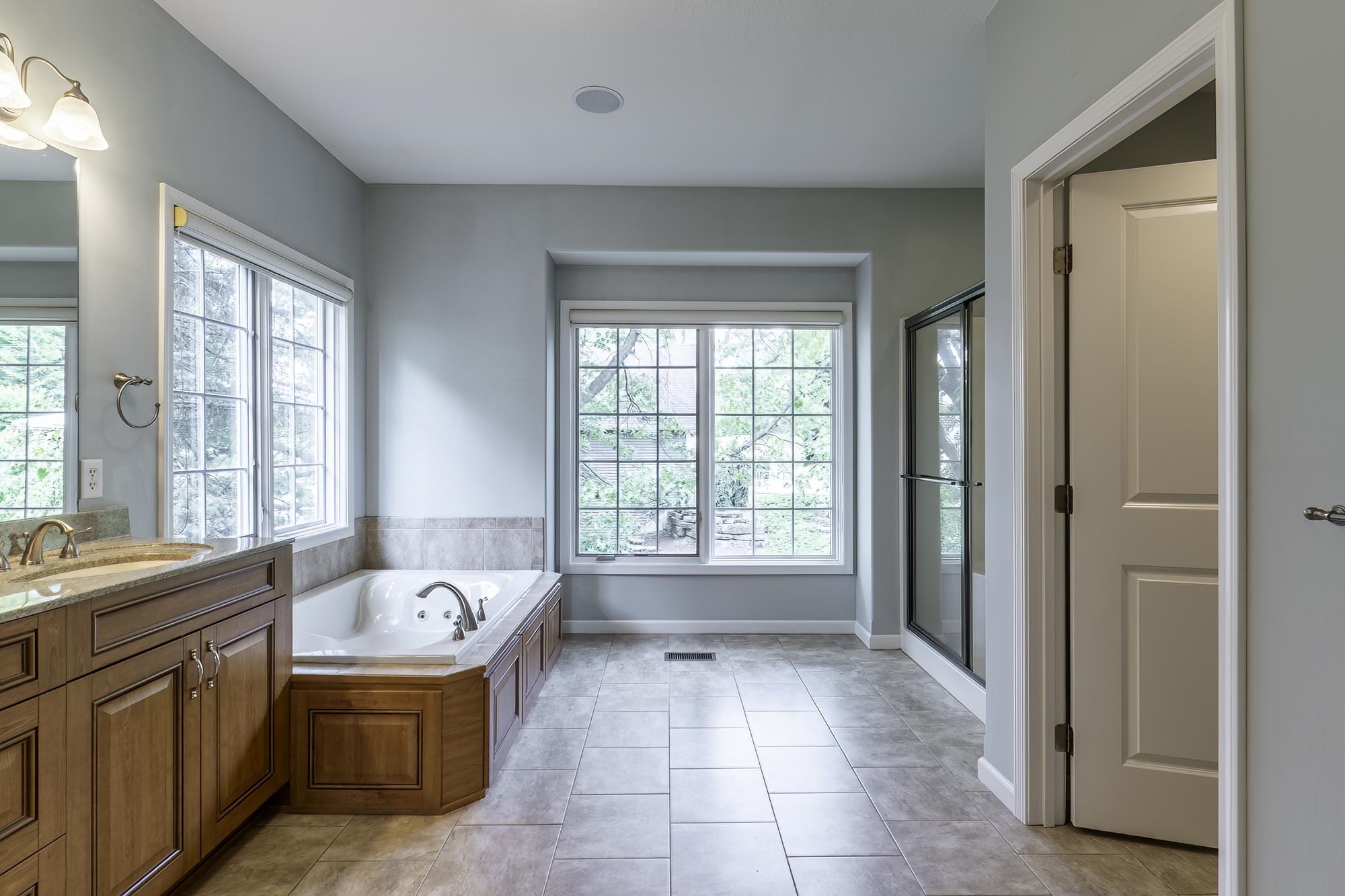 Neutral-toned bathroom featuring a built-in jetted tub with wood-finish paneling, a large window with grid panes, a standalone shower with a glass enclosure, a single vanity with wood-finish cabinetry, and tiled flooring