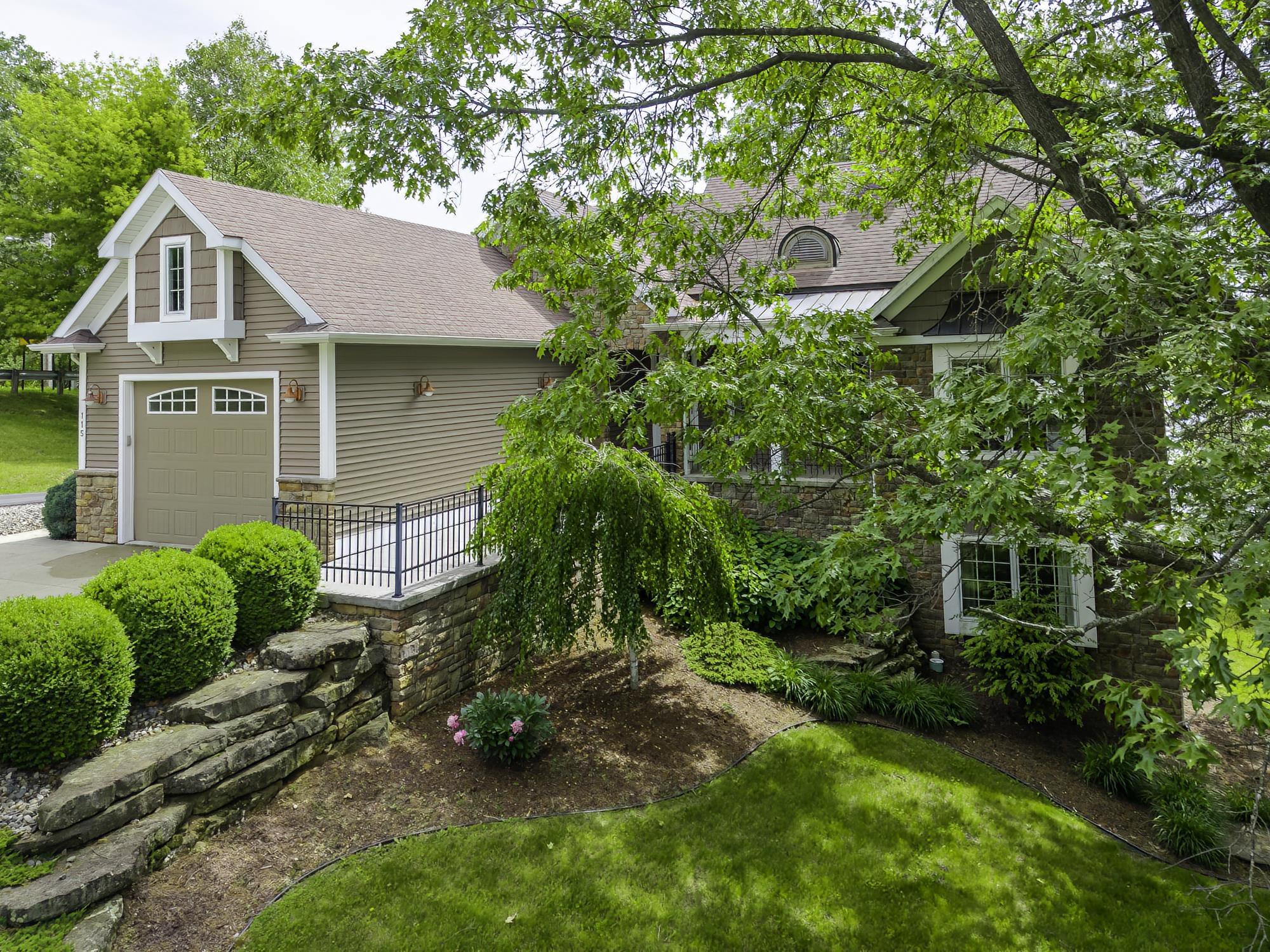 Exterior featuring stone and siding finishes, an attached garage with a dormer, mature trees, and landscaped grounds