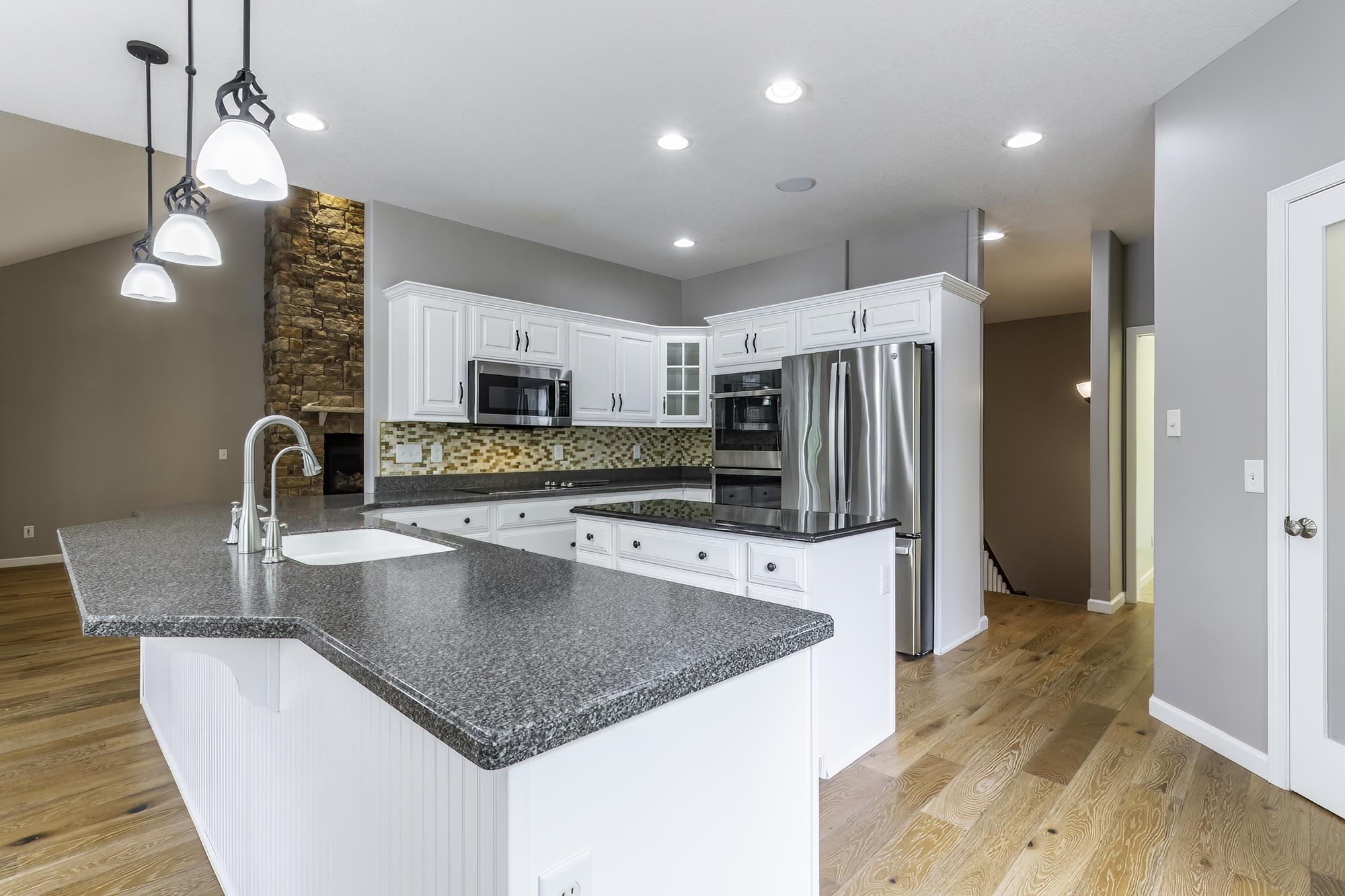 Open-concept kitchen featuring an island with a sink and pendant lighting, white cabinetry, integrated stainless steel appliances, a stone-finish backsplash, and light wood-finish flooring