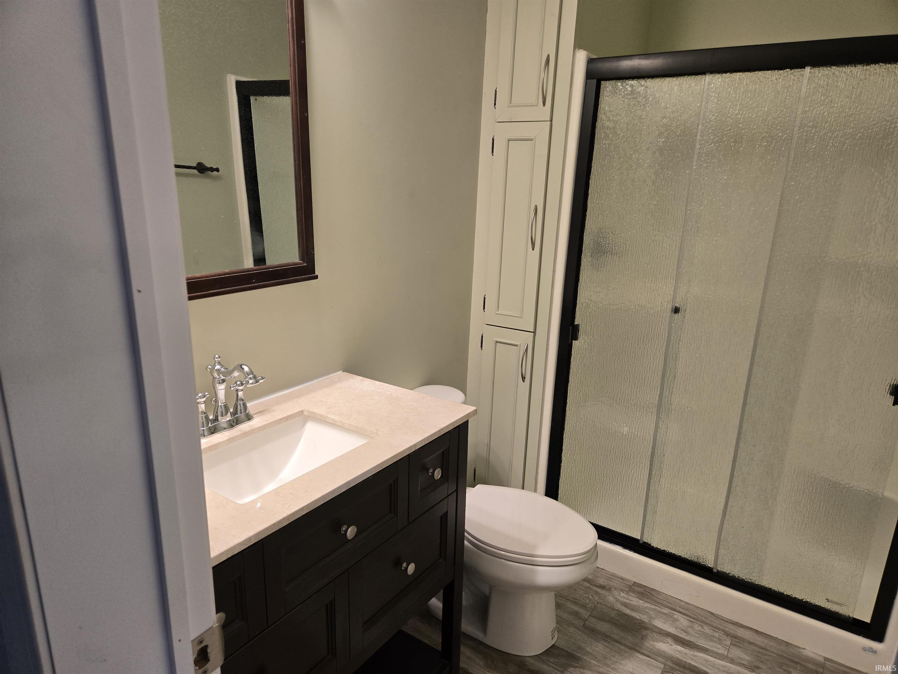 Bathroom featuring a dark wood vanity with a light-colored countertop and integrated sink, a framed mirror, a shower with textured glass doors and a black frame, light wood-finish flooring, and a built-in storage cabinet
