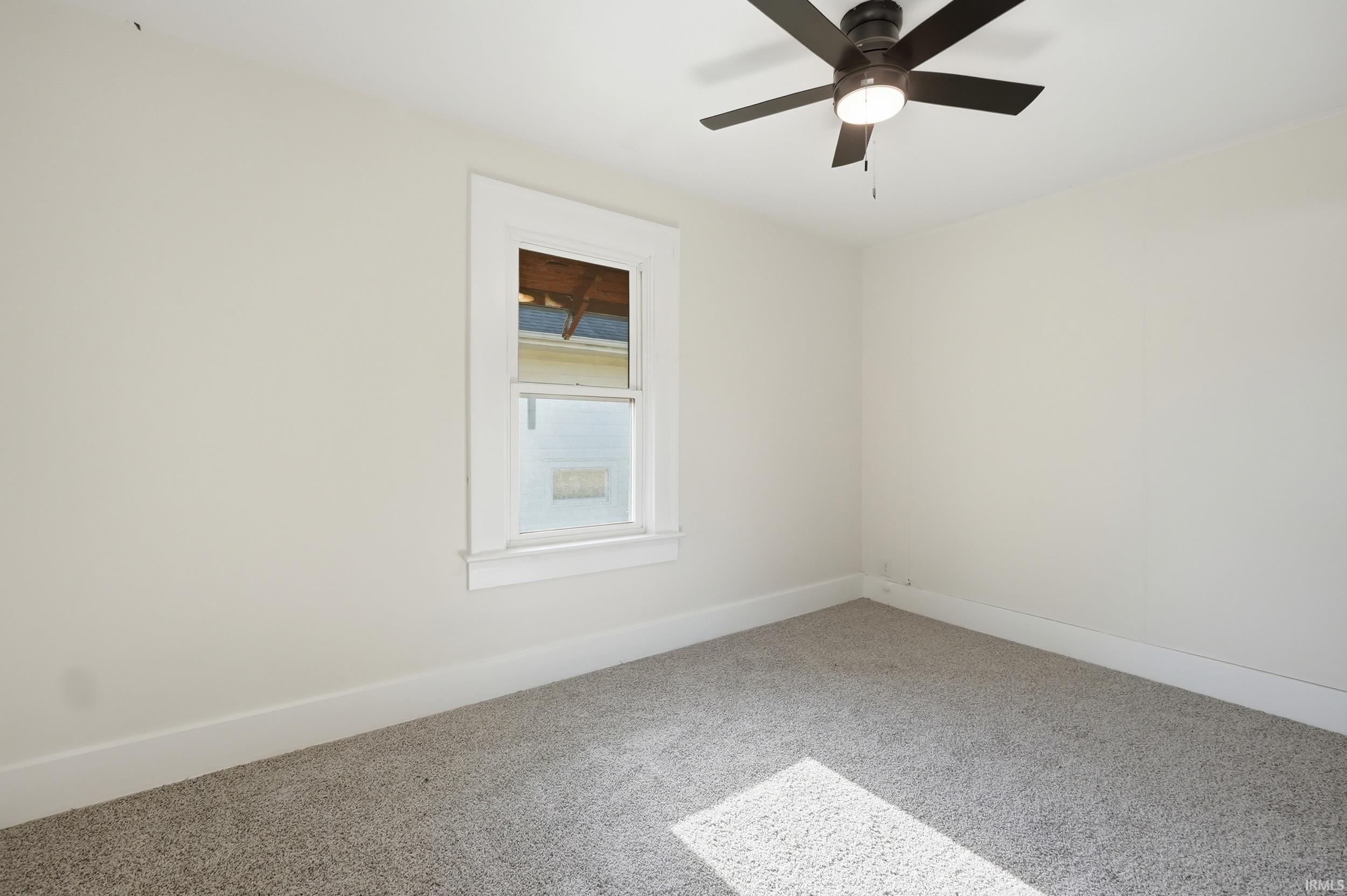 Carpeted interior space featuring a window with white trim, a dark-toned ceiling fan with integrated lighting, and white baseboard molding