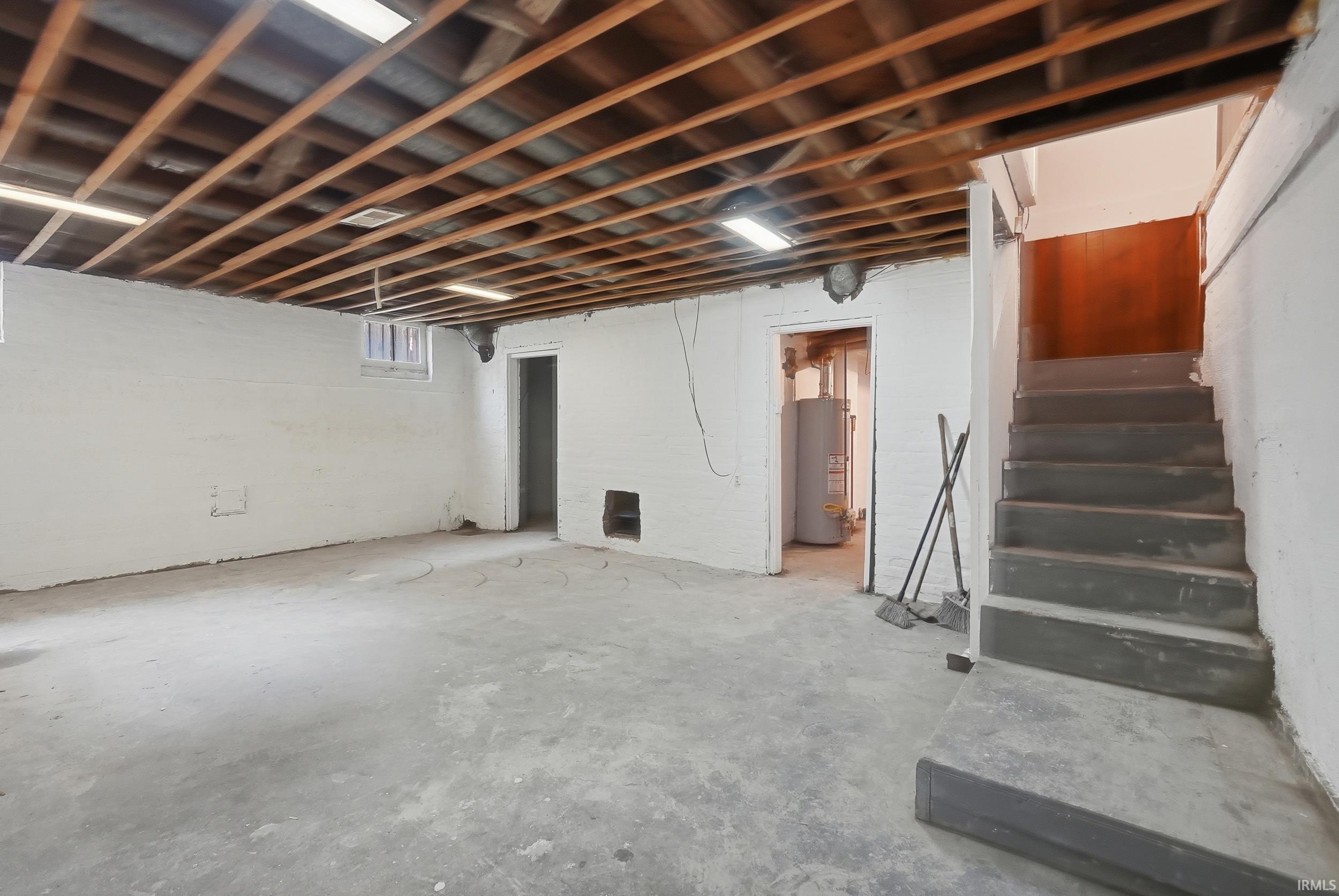 Unfinished basement featuring exposed wood ceiling joists, white painted brick walls, concrete flooring, and a concrete staircase