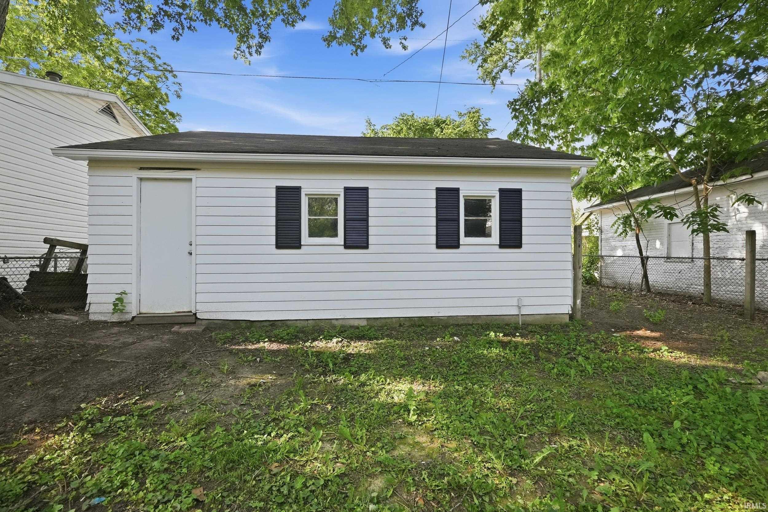 Detached garage featuring white vinyl siding, black shutters, two windows, a single entry door, and a dark shingle roof