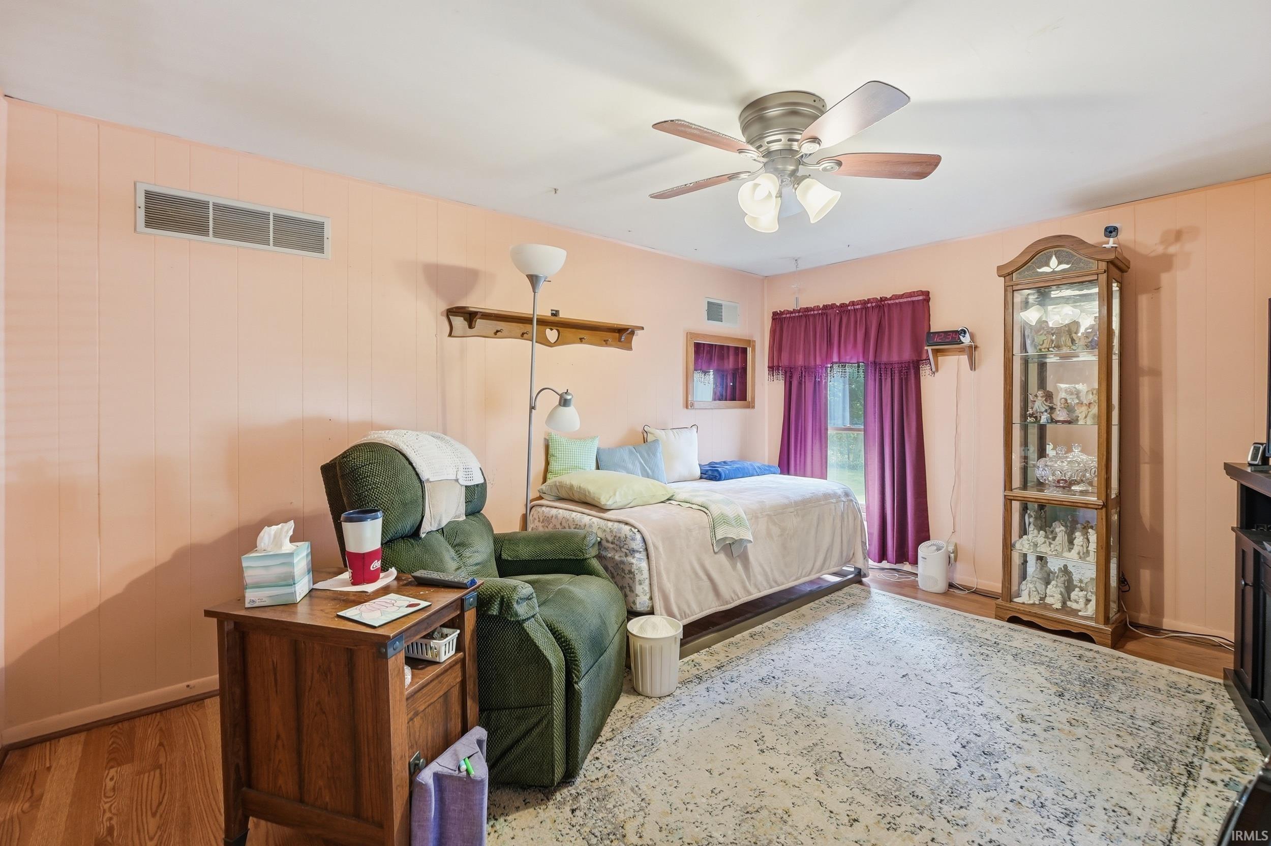 Spacious room featuring wood-finish flooring, a ceiling fan with integrated lighting, and a large window with privacy curtains