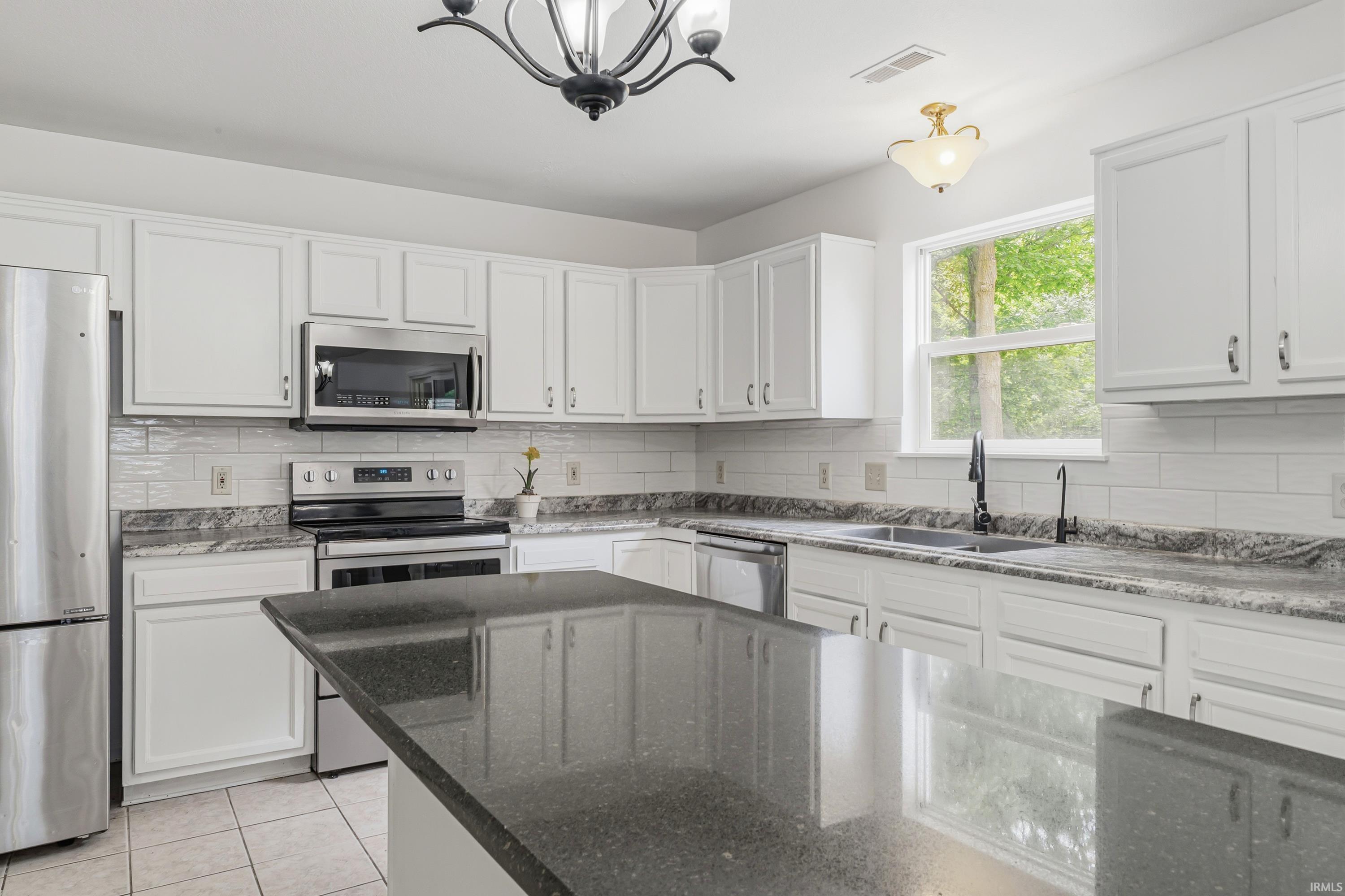 Well-appointed kitchen featuring white cabinetry, dark stone countertops, stainless steel appliances, a tile backsplash, and a central island with an integrated sink