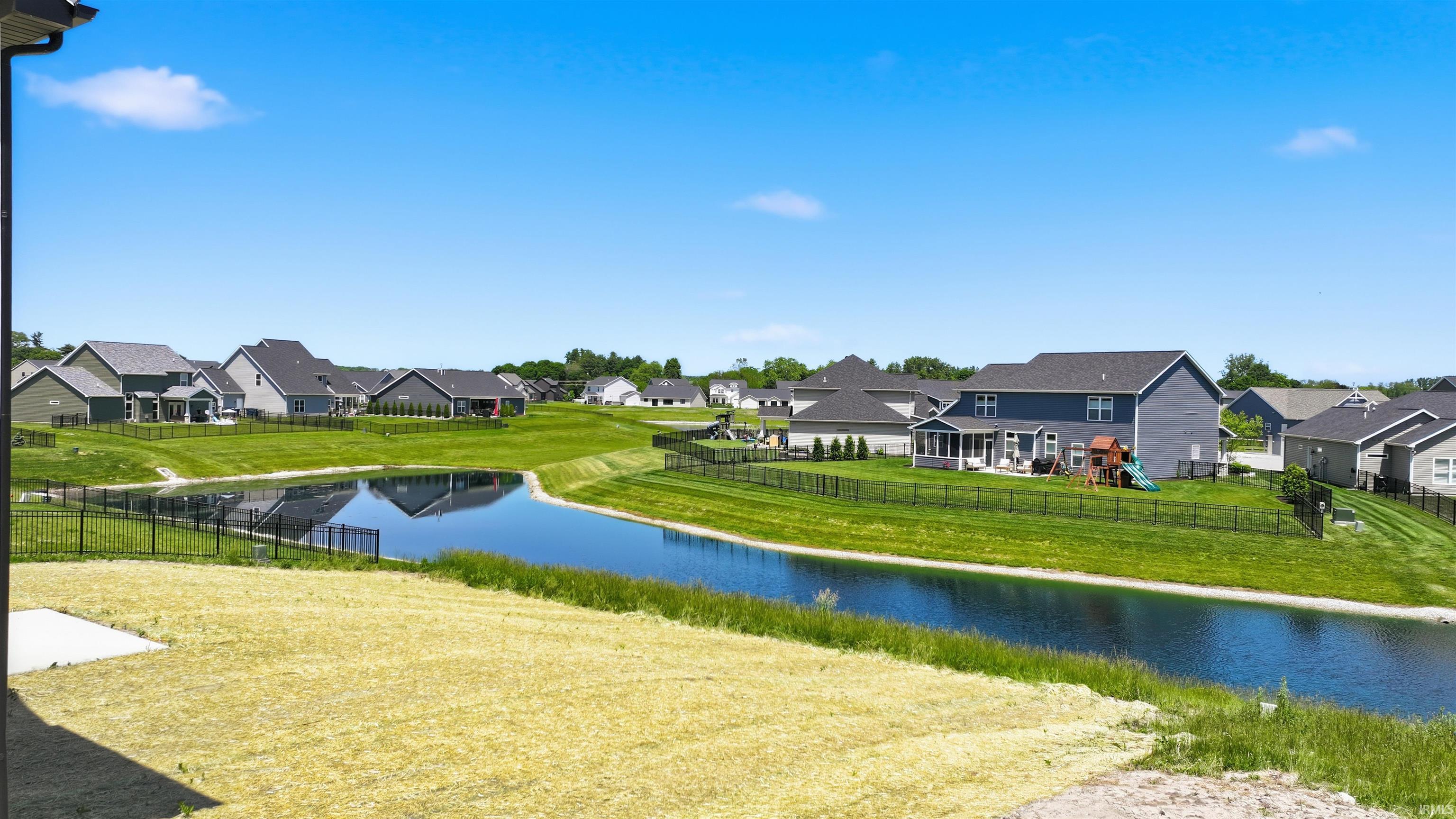 Expansive backyard featuring a pond with surrounding green space, a black metal fence, and a concrete patio pad