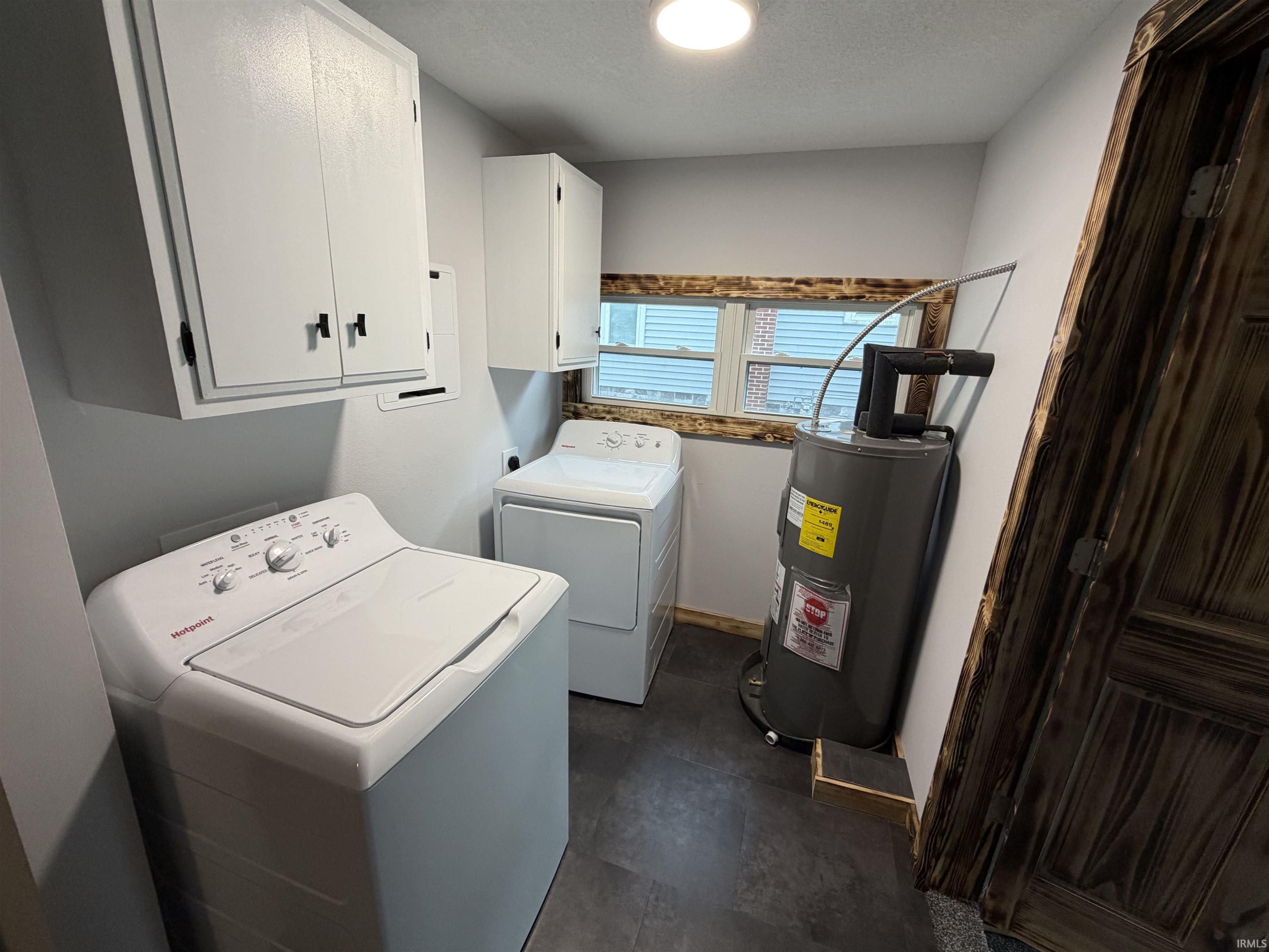 Dedicated laundry area featuring white wall cabinetry, a window with wood trim, a water heater, and dark tile flooring