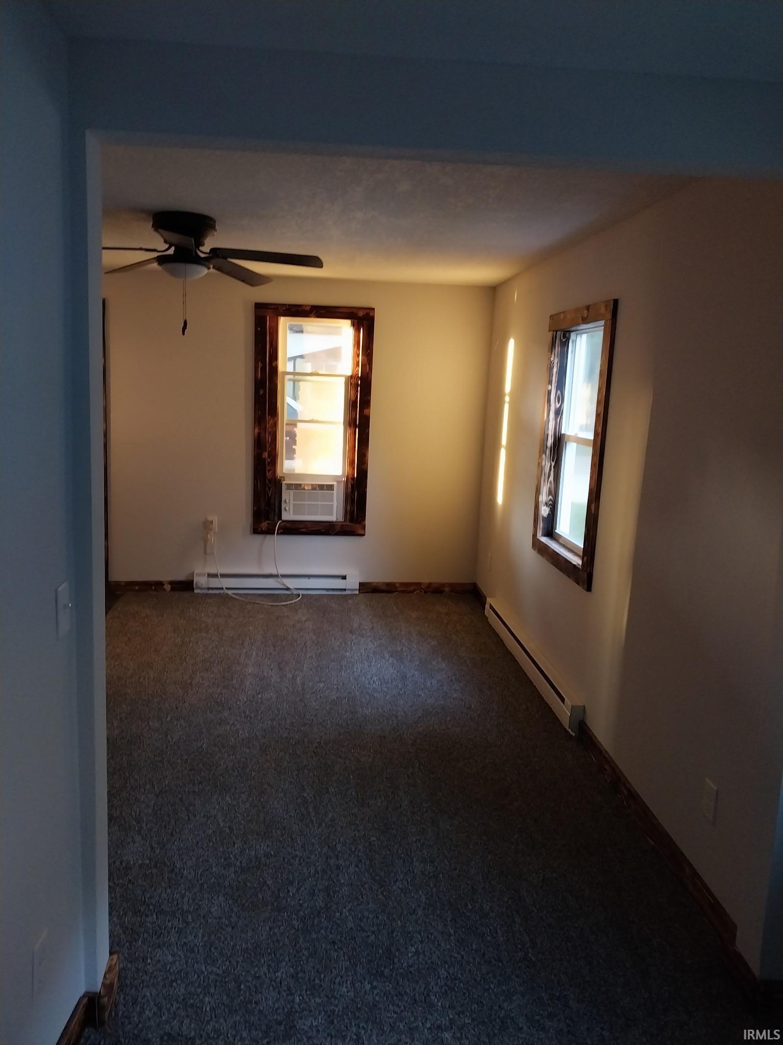 Carpeted interior space featuring two windows with wood trim, a ceiling fan with light fixture, and baseboard heating