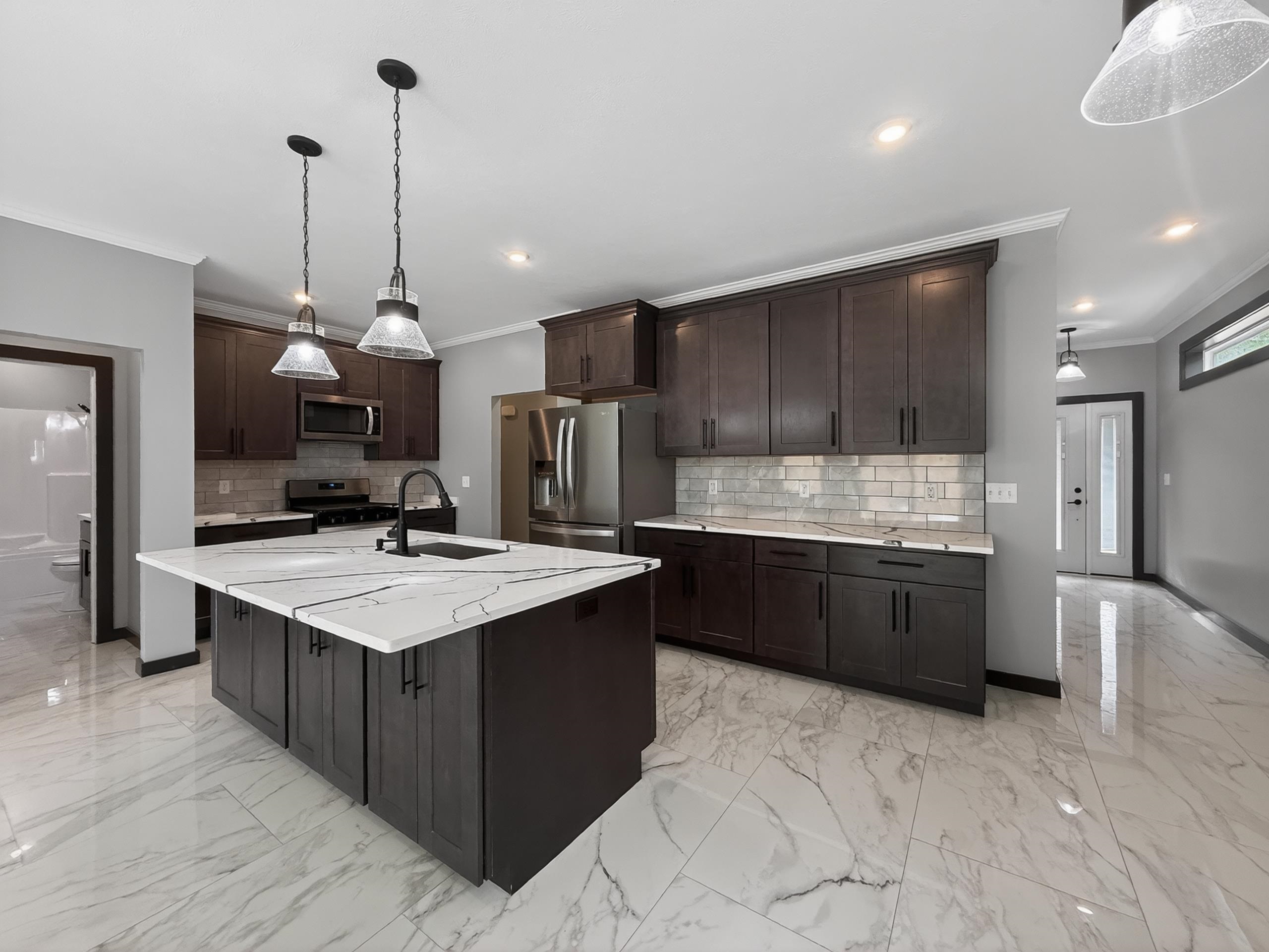 Expansive kitchen featuring a large central island with a white veined countertop, dark wood-finish cabinetry, subway tile backsplash, and stainless steel appliances