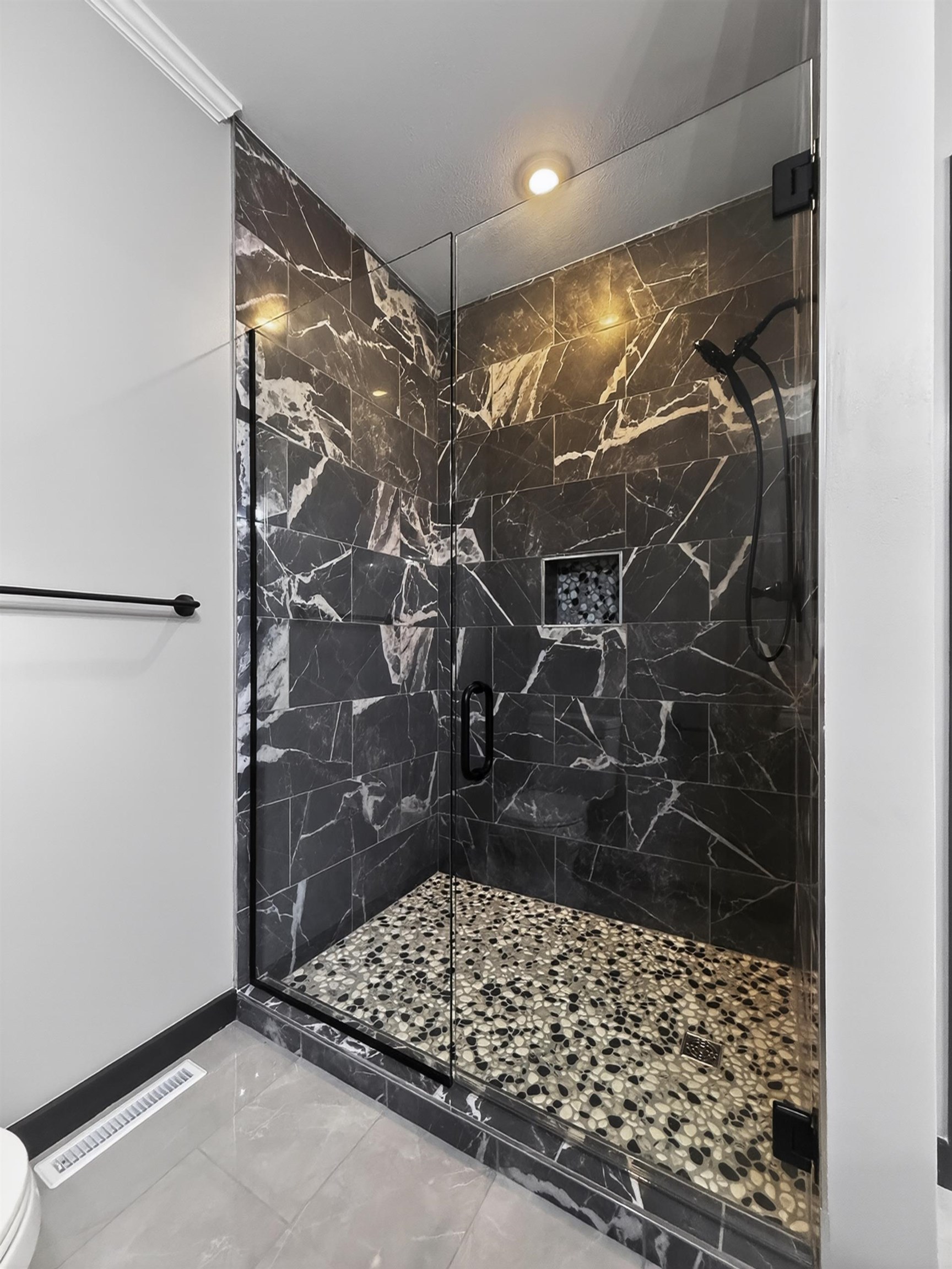 Frameless glass shower enclosure featuring black marble-style tile walls, a built-in shower niche, and a pebble-style floor