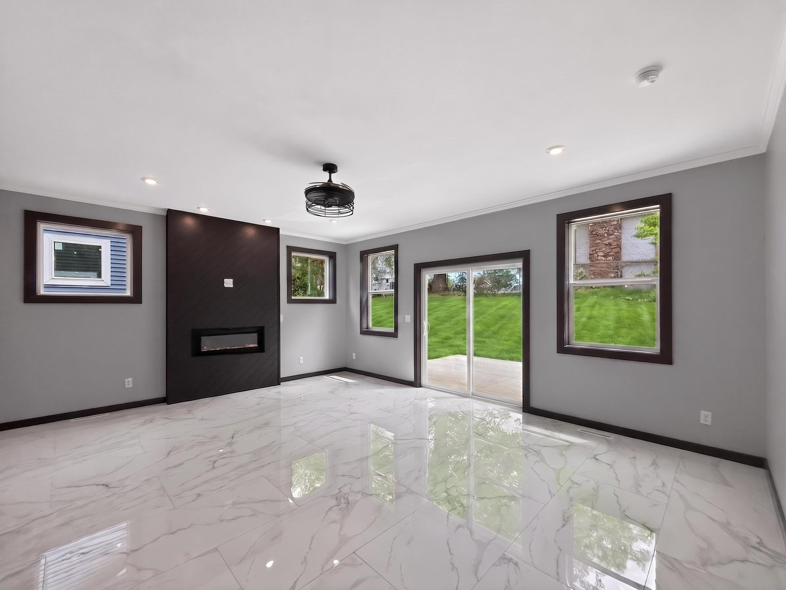 Spacious room featuring glossy marble-patterned flooring, a modern electric fireplace insert within a dark accent wall, recessed ceiling lighting, a contemporary ceiling fan fixture, and sliding glass doors opening to the backyard