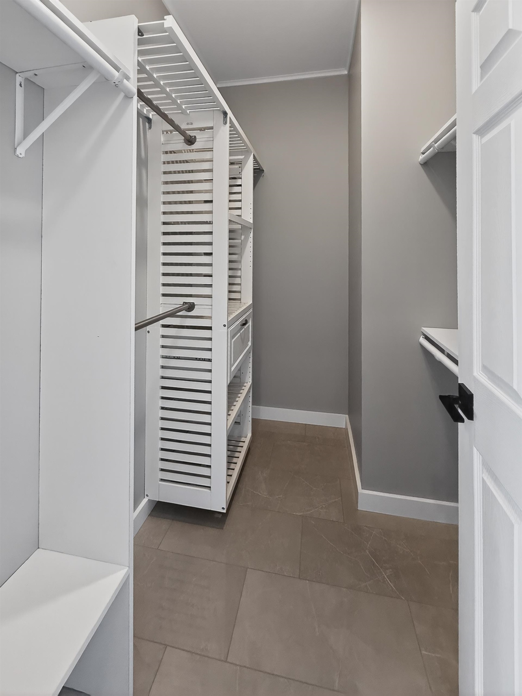 Walk-in closet featuring light gray walls, built-in white shelving, multiple hanging rods, and large format tile flooring