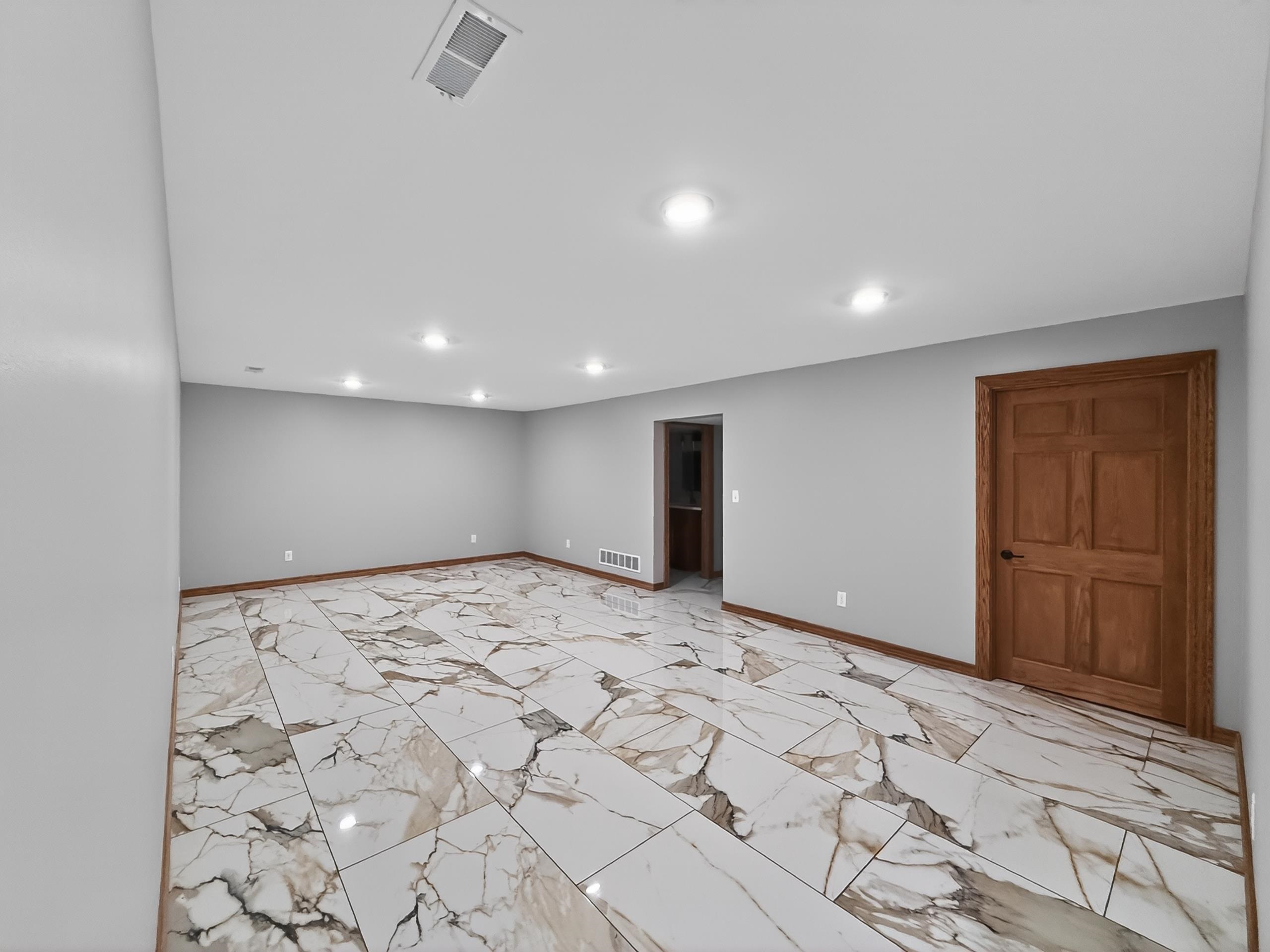Spacious room featuring large format tile flooring with a marble-like pattern, light gray wall paint, recessed ceiling lighting, a wood-paneled door with dark hardware, and coordinating wood baseboards