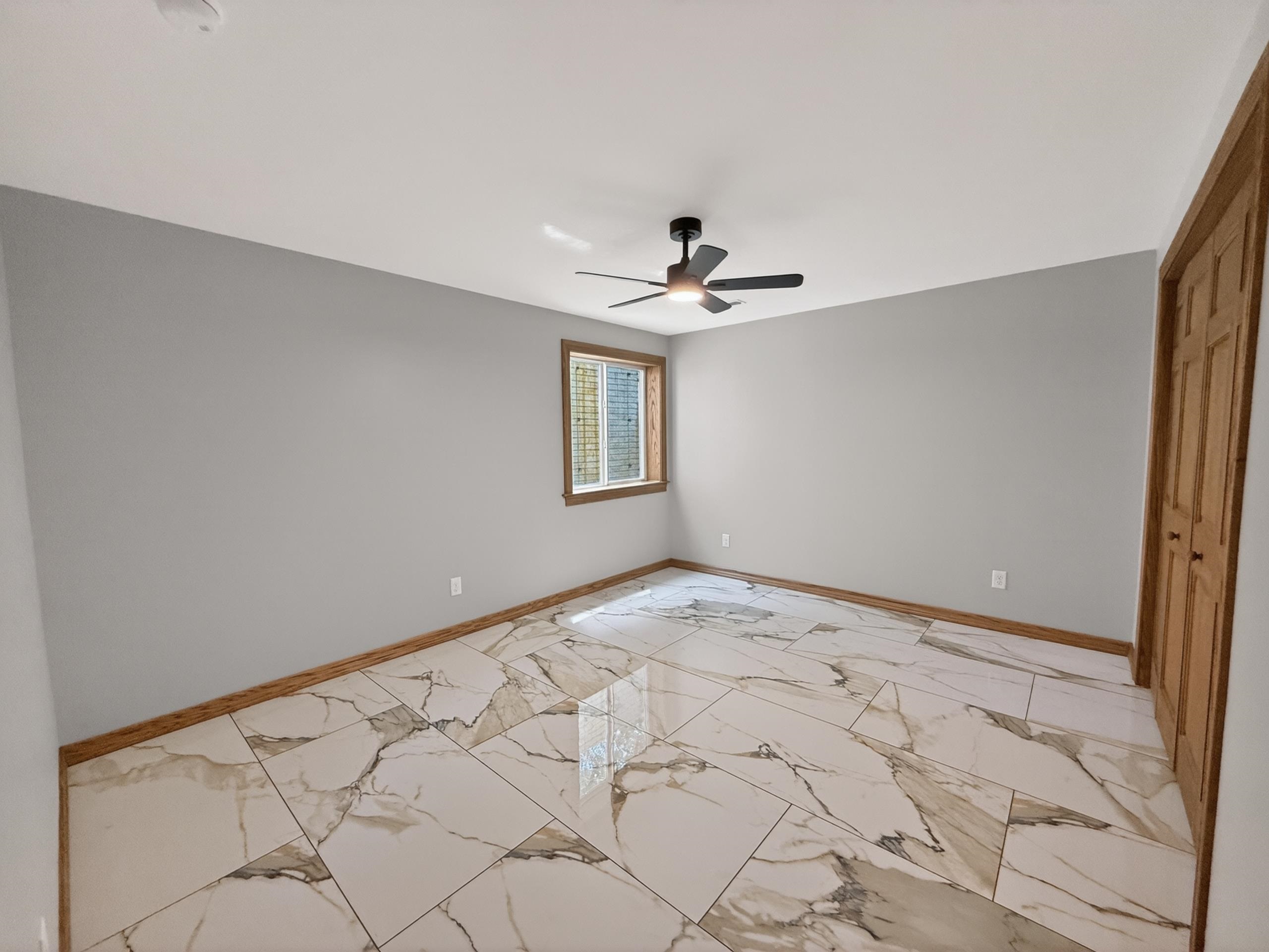 Spacious room featuring large format tile flooring with a marble-effect pattern, light gray walls, a modern ceiling fan, wood trim, and a window with blinds