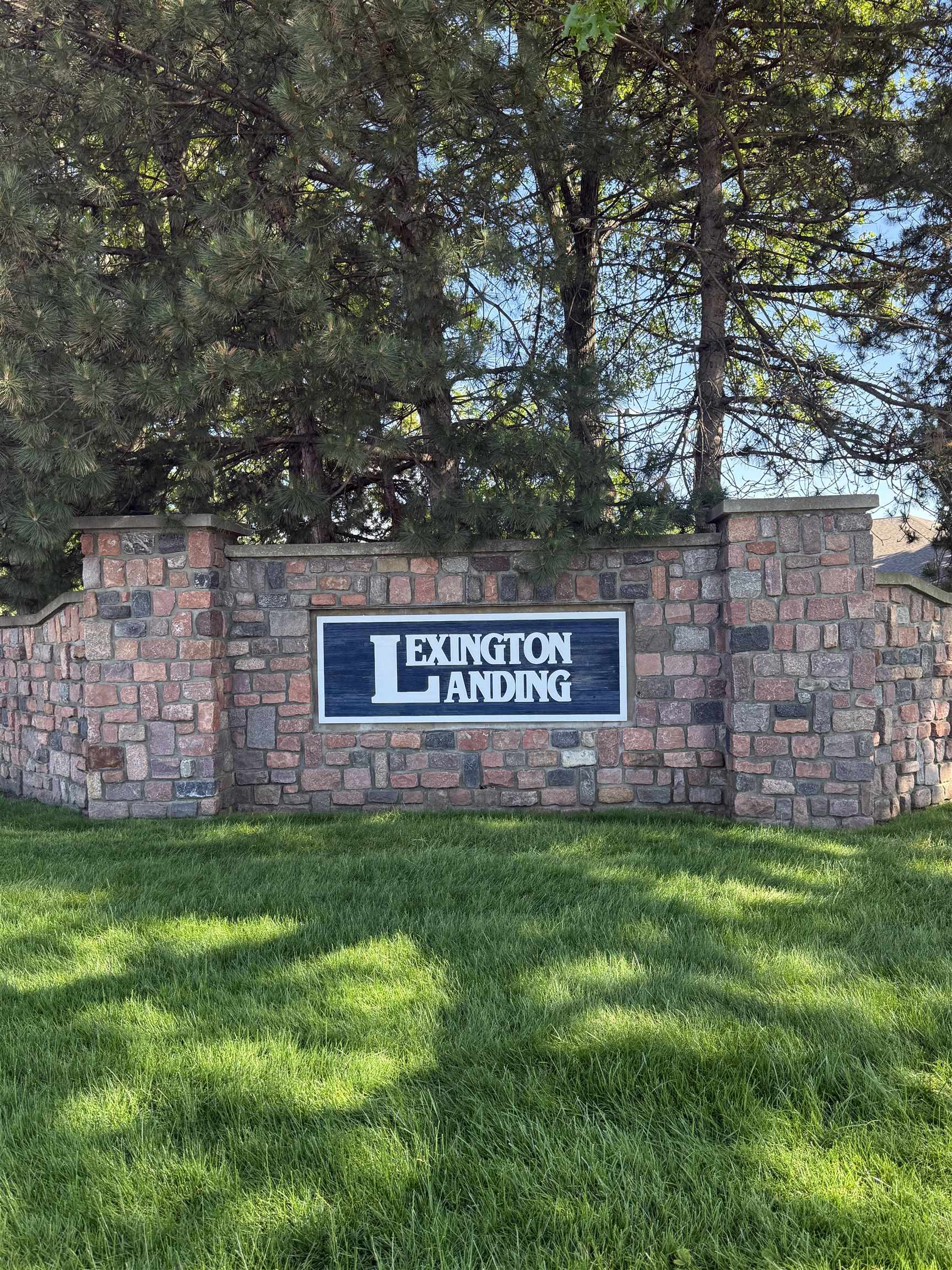 Lexington Landing community entrance featuring a stone wall, integrated signage, mature evergreen trees, and a green lawn