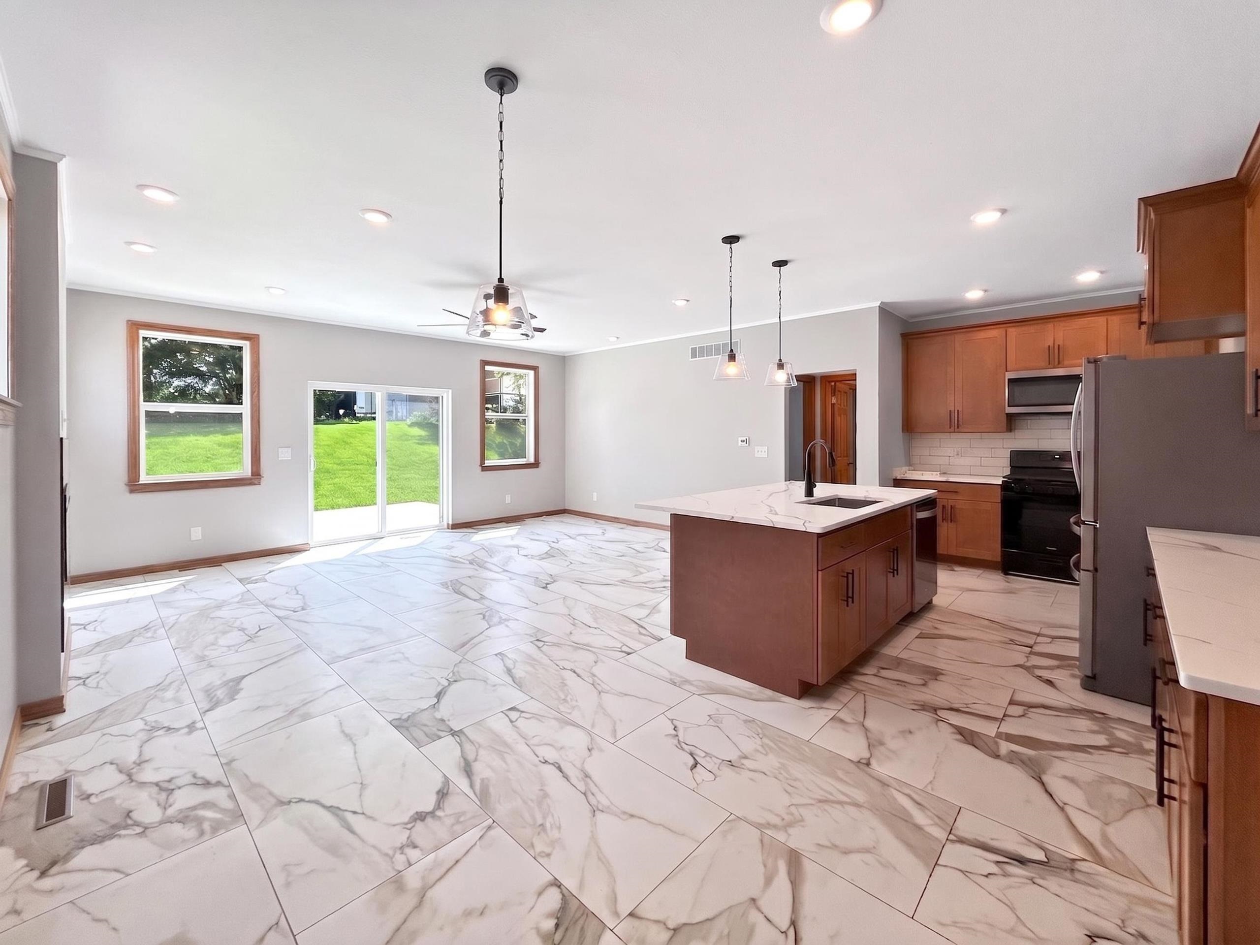 Spacious open-concept kitchen and living area featuring large format white and gray veined tile flooring, a kitchen island with a built-in sink and pendant lighting, wood cabinetry with white countertops, and a full suite of black kitchen appliances