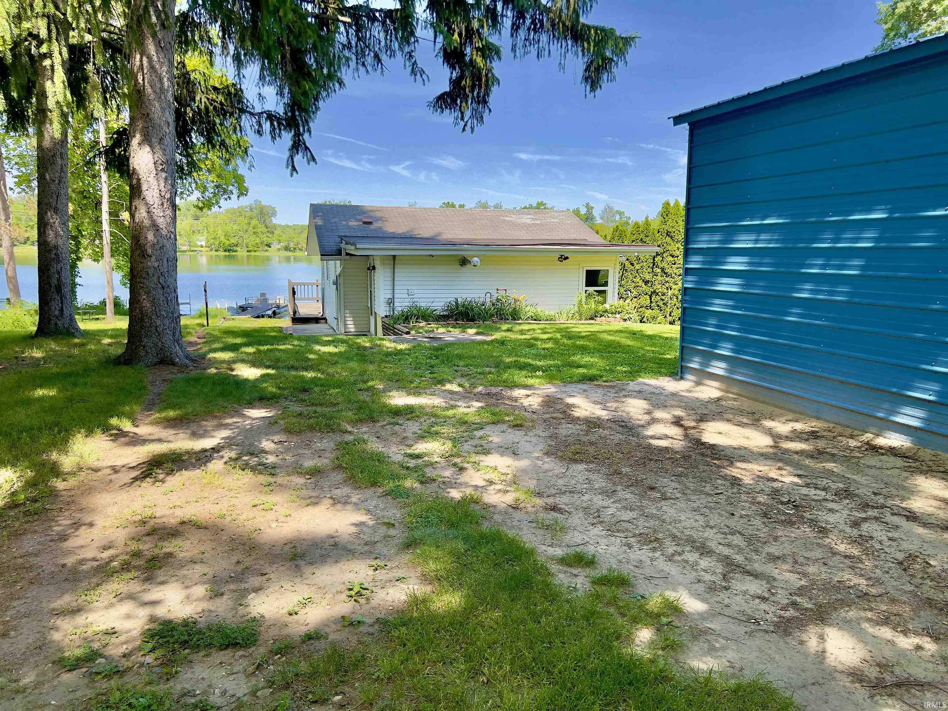 Waterfront property featuring a white siding structure with a gray shingle roof, a blue-sided outbuilding, a green lawn, and mature trees