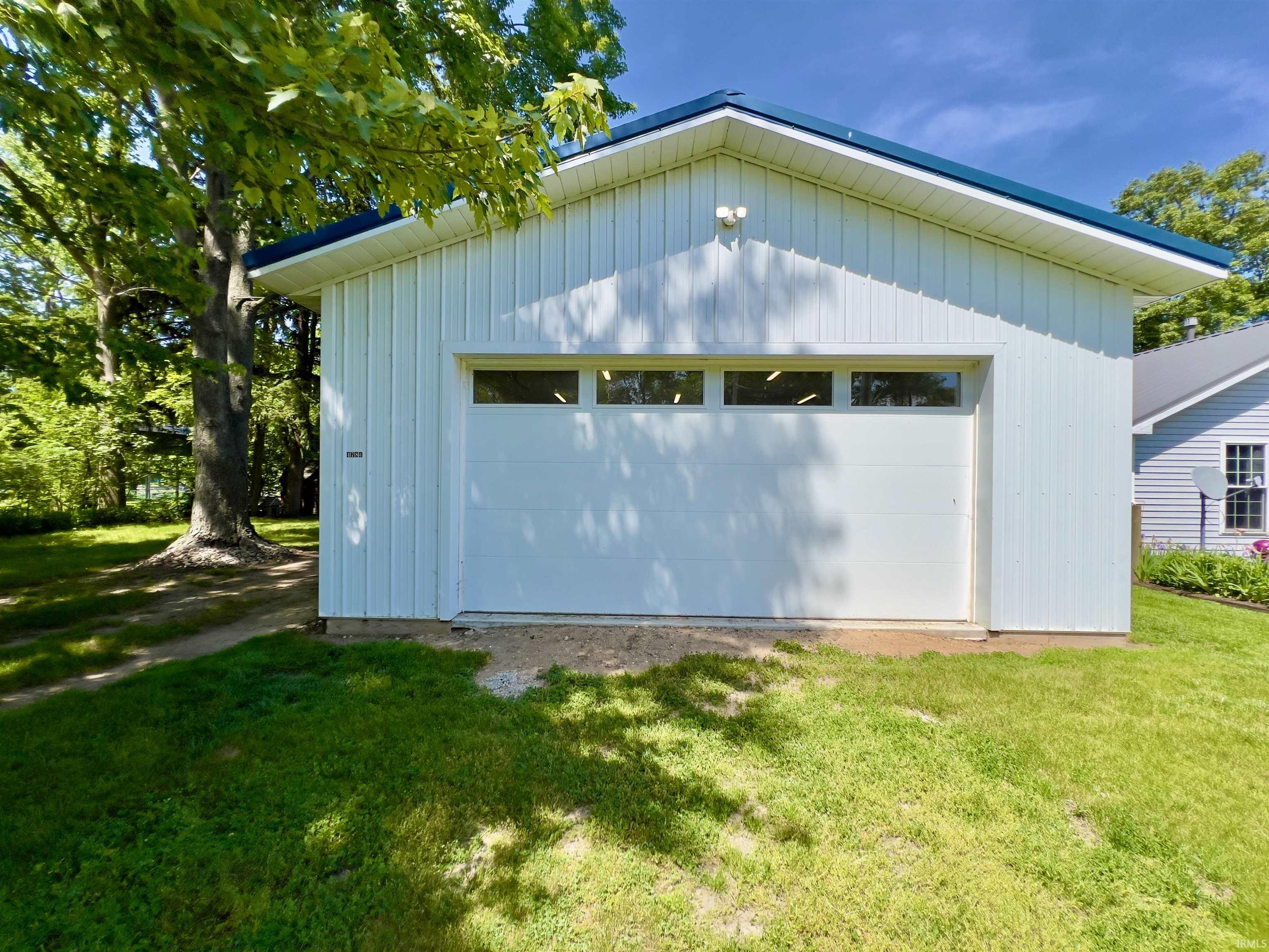 Detached outbuilding featuring white vertical siding, a white garage door with horizontal transom windows, a blue metal roof, and exterior wall-mounted lighting