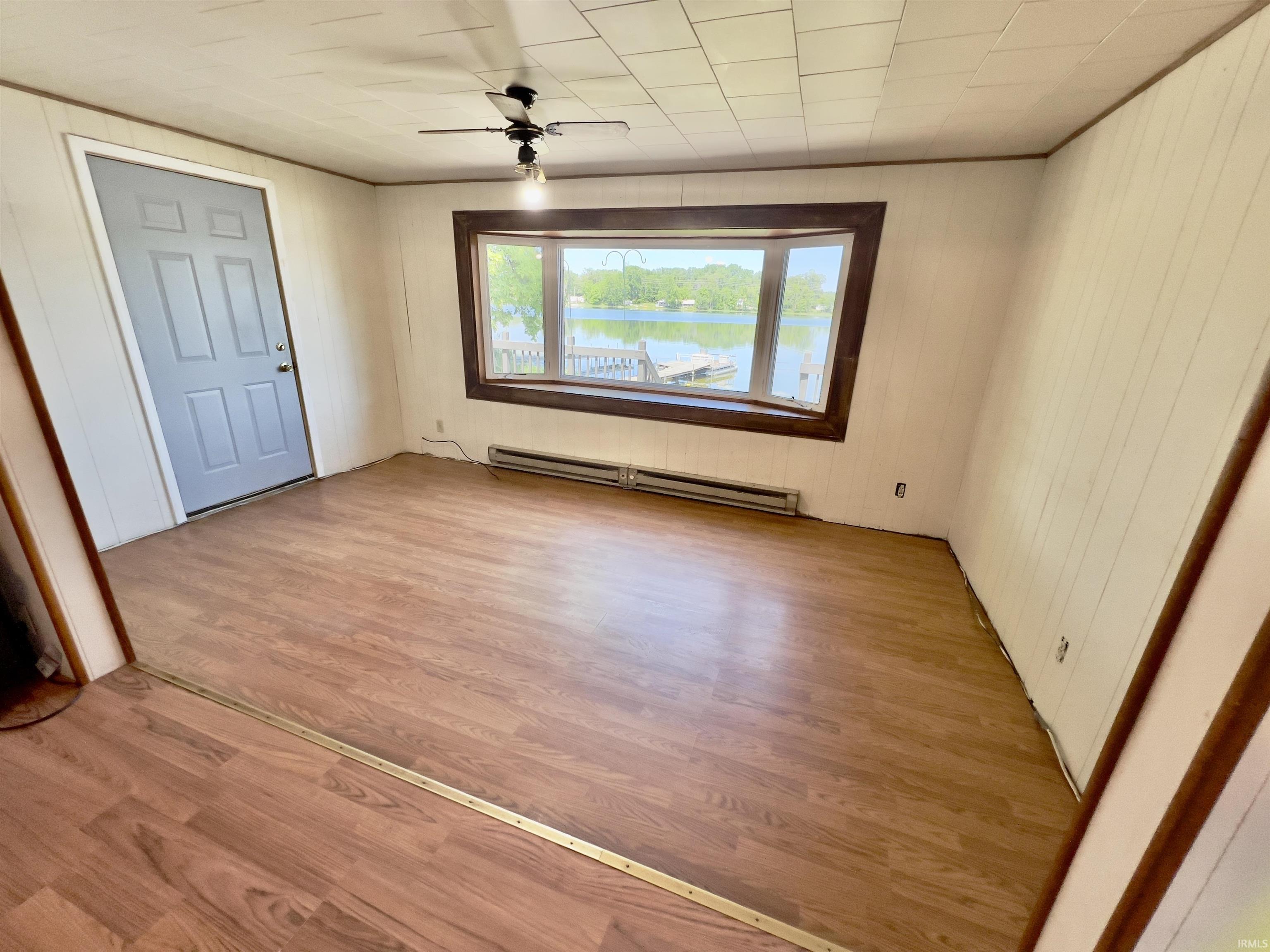 Room featuring bay window, water views, wood-finish flooring, white paneling, a ceiling fan, and a light gray entry door