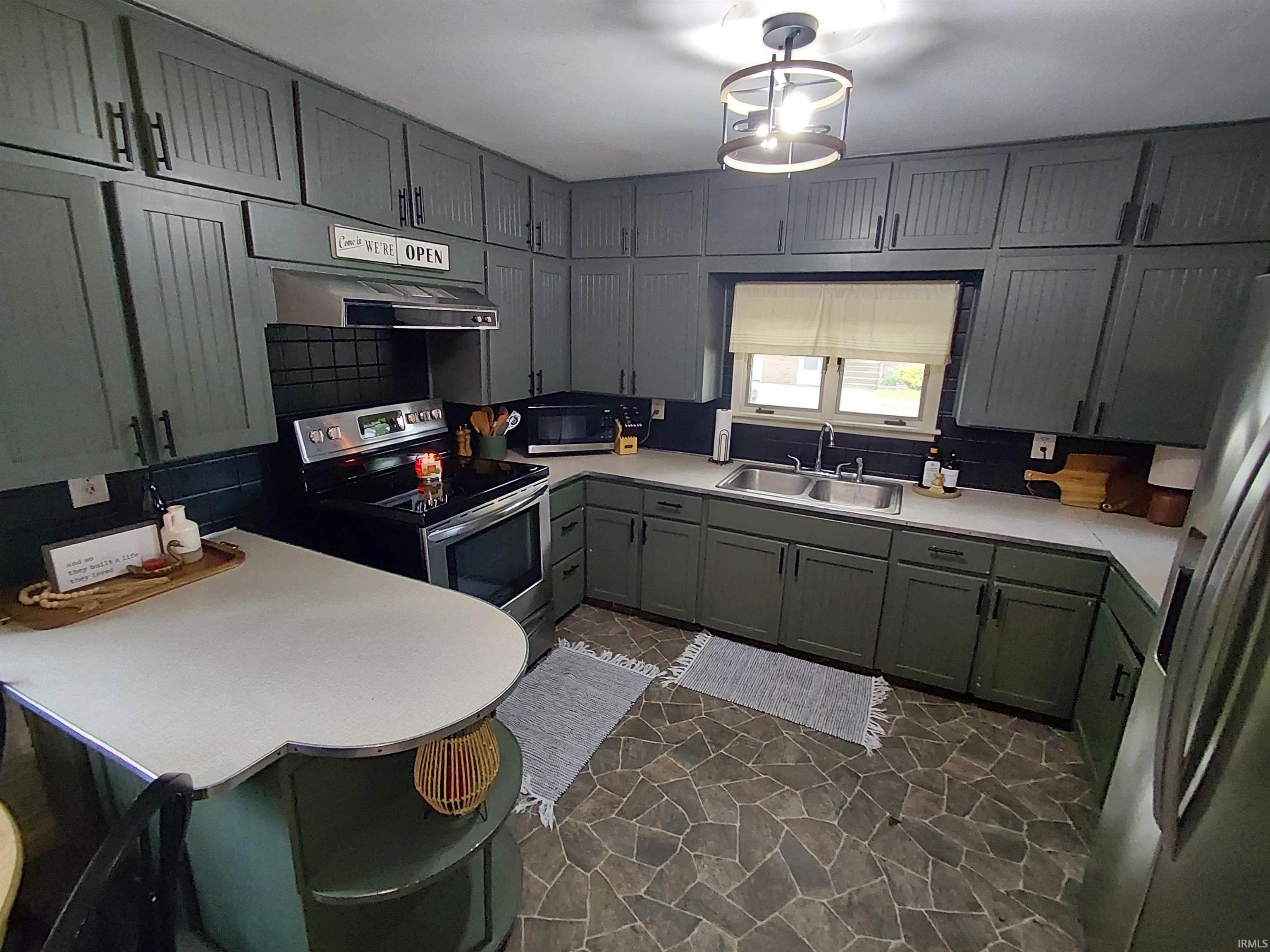 Kitchen featuring dark green cabinetry with black hardware, stainless steel appliances, a double basin sink, light countertops, and a dark tile backsplash
