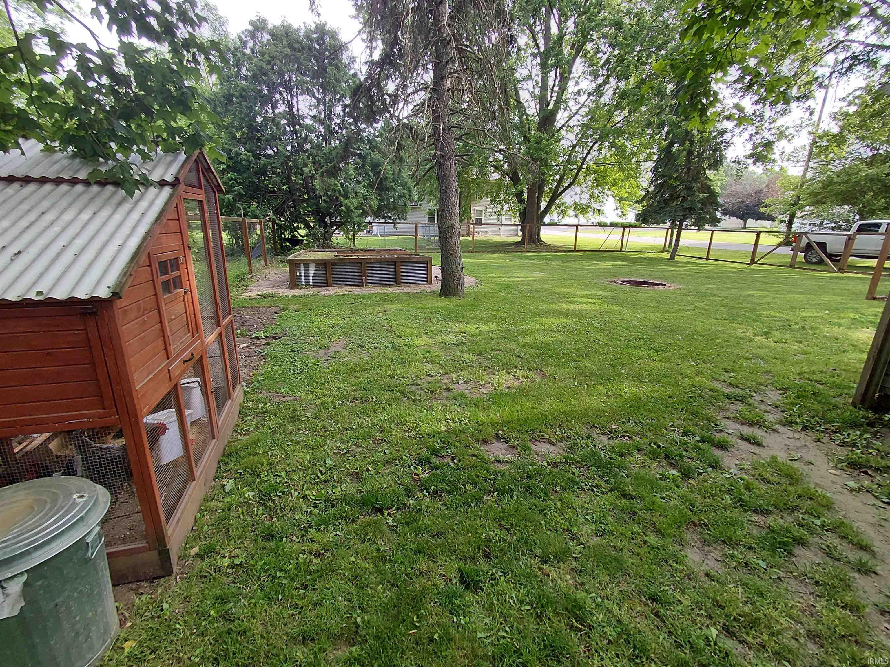 Expansive grassy yard featuring a wooden coop with corrugated roof, a raised garden bed, a fire pit, and mature trees