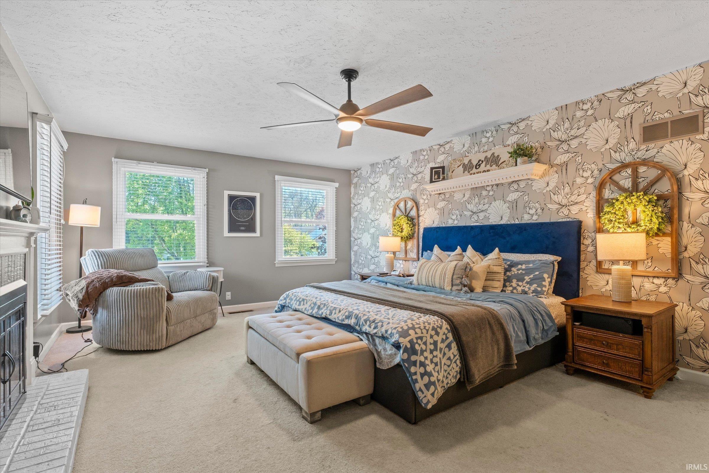 Bedroom featuring a fireplace with a light-colored brick hearth, two large windows, a ceiling fan with integrated lighting, and a feature wall with botanical pattern wallpaper
